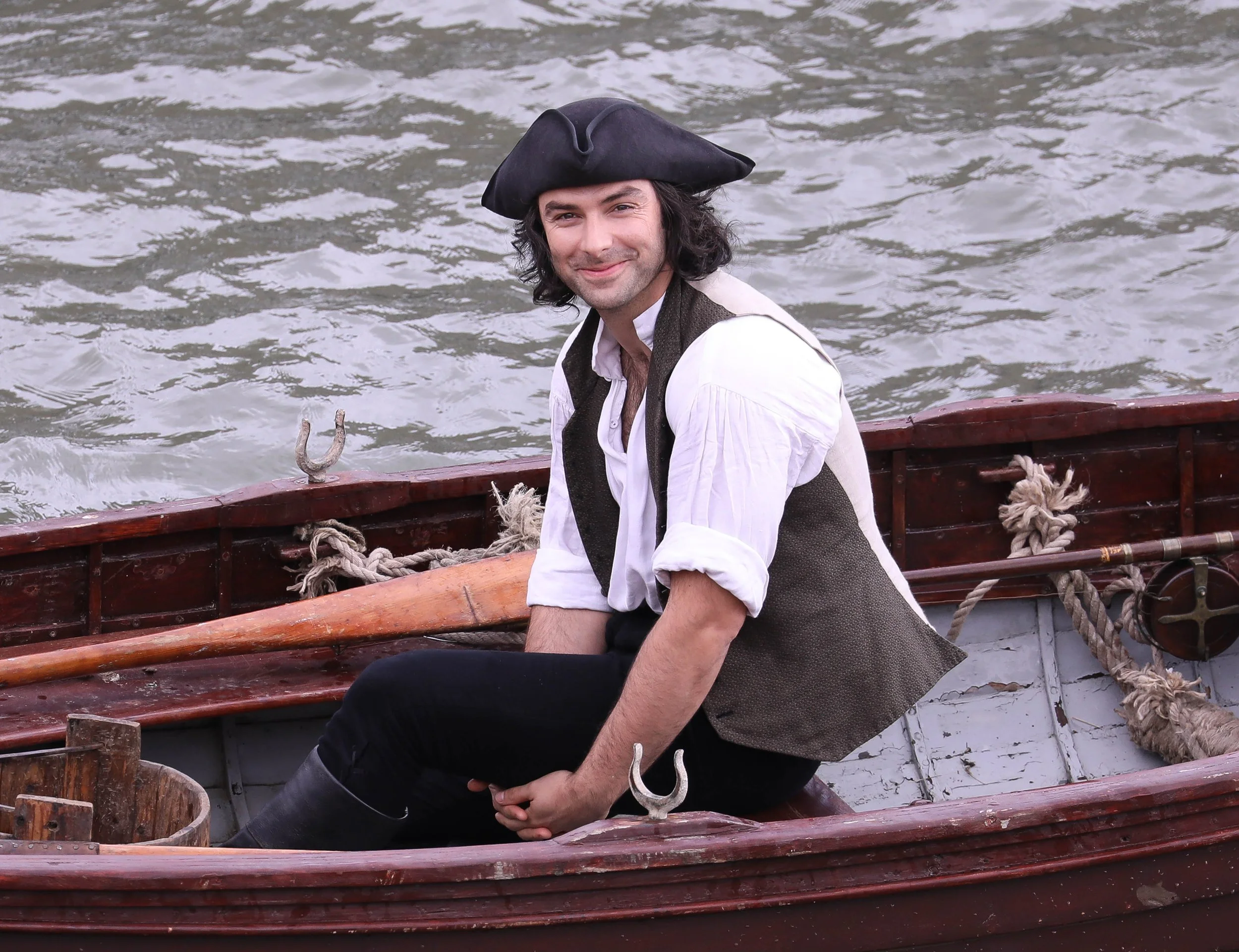 A man dressed as a pirate sitting on a wooden boat in the water, wearing a black pirate hat, a white shirt, and a vest, smiling at the camera.