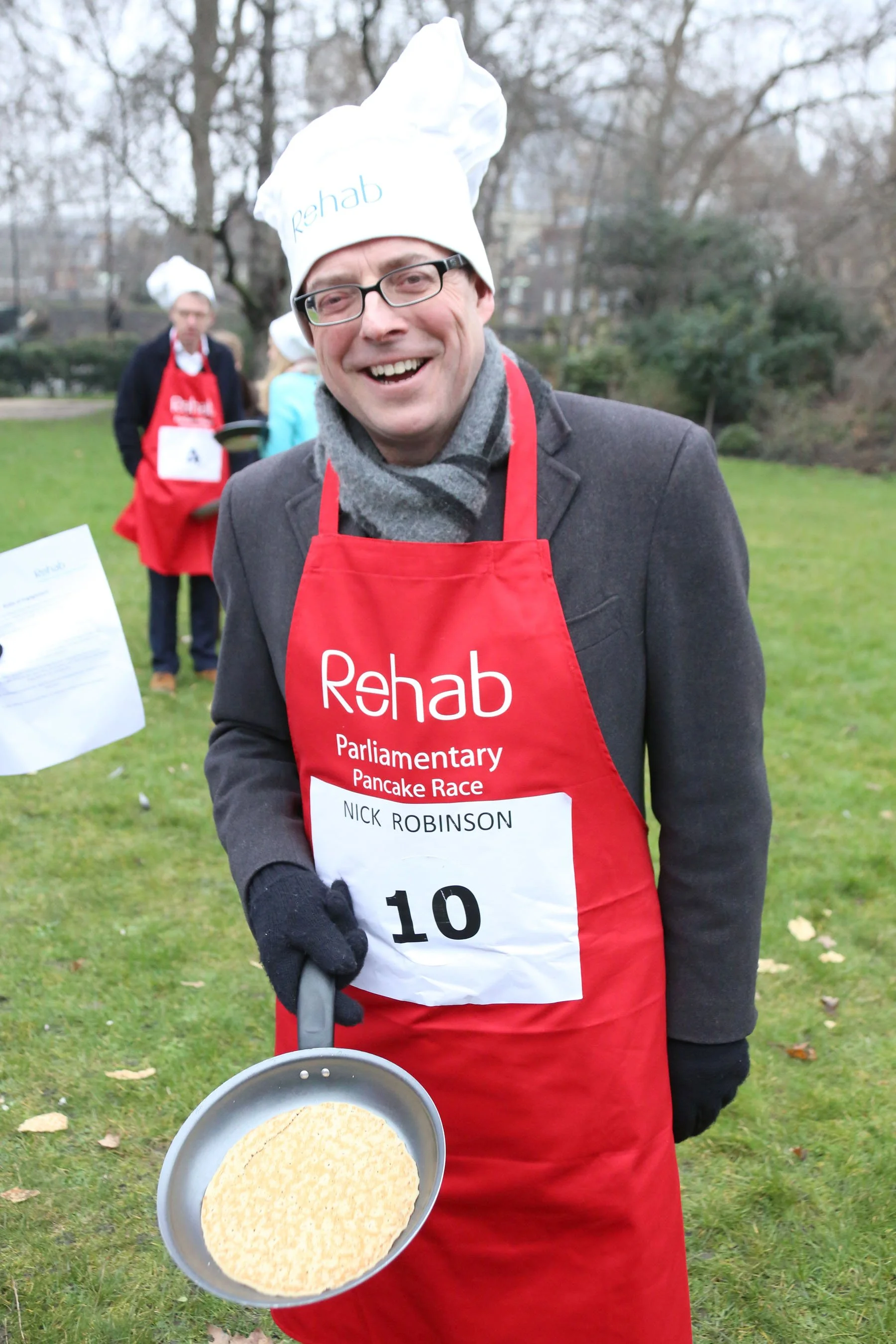 A man wearing a red apron and a chef's hat that reads 'Rehab' is smiling outside during a pancake race event. He is holding a frying pan with a pancake in it. Other people in aprons and chef hats are in the background, and the setting appears to be a