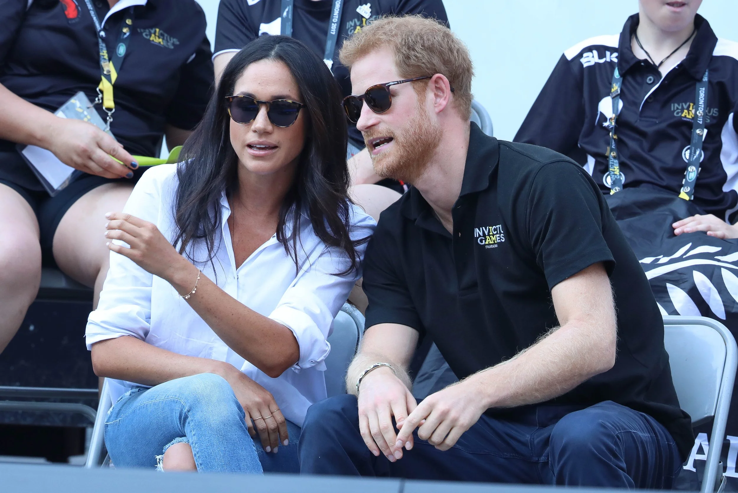 Prince Harry and Meghan Markle sitting together in the stands at an event, engaging in conversation, with other attendees nearby.
