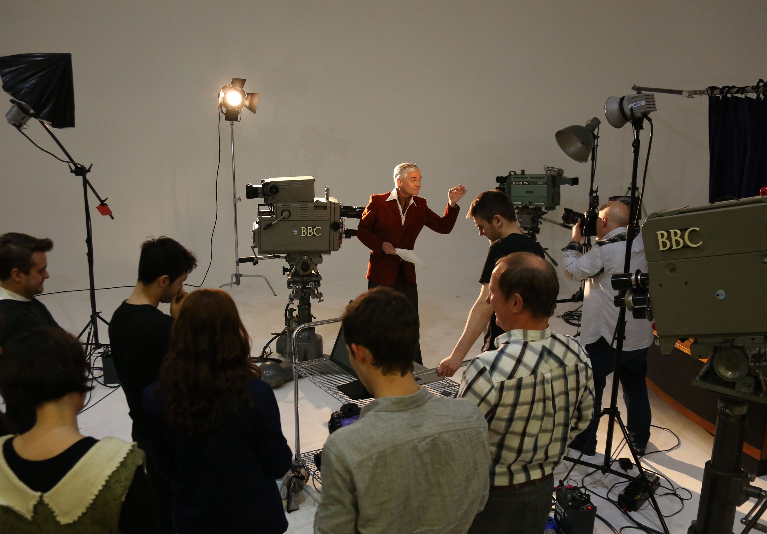 A woman with short, gray hair in a red jacket is speaking or presenting to an audience in a studio with professional camera equipment and lighting.