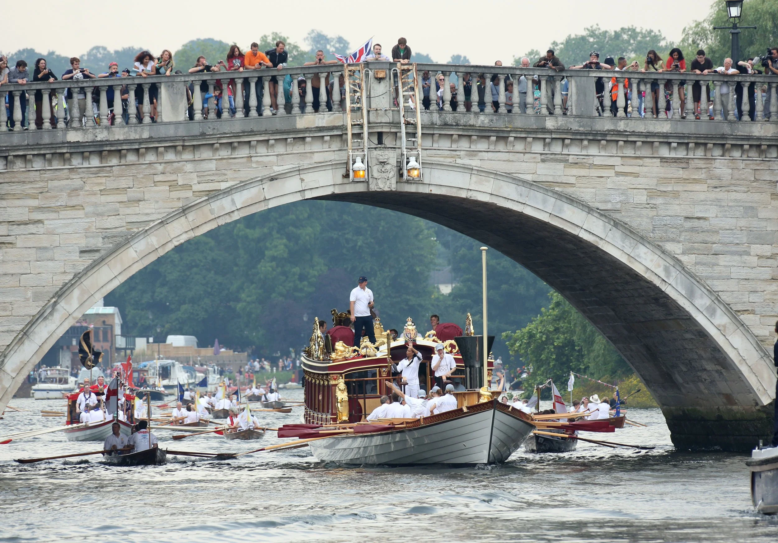 A parade of decorated boats with people on board, passing under a stone bridge on a river, with spectators on the bridge and along the riverbanks.