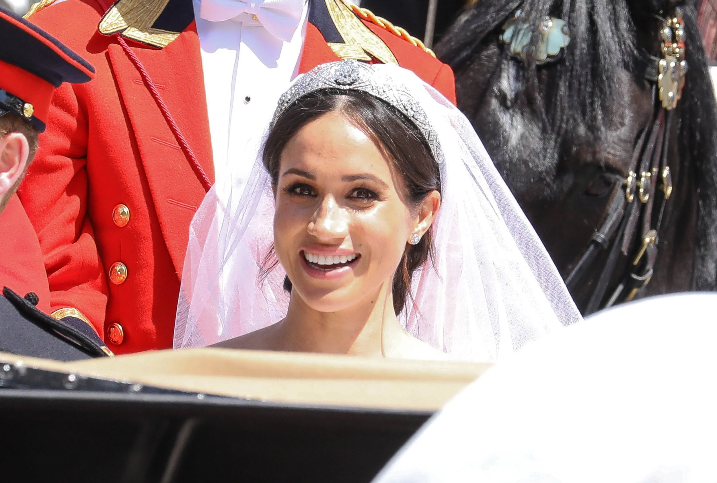 A bride with dark hair smiling, wearing a white veil with a decorative headpiece, in a formal wedding setting, a man in a red military uniform stands beside her, with a person in a dark outfit and horse in the background.