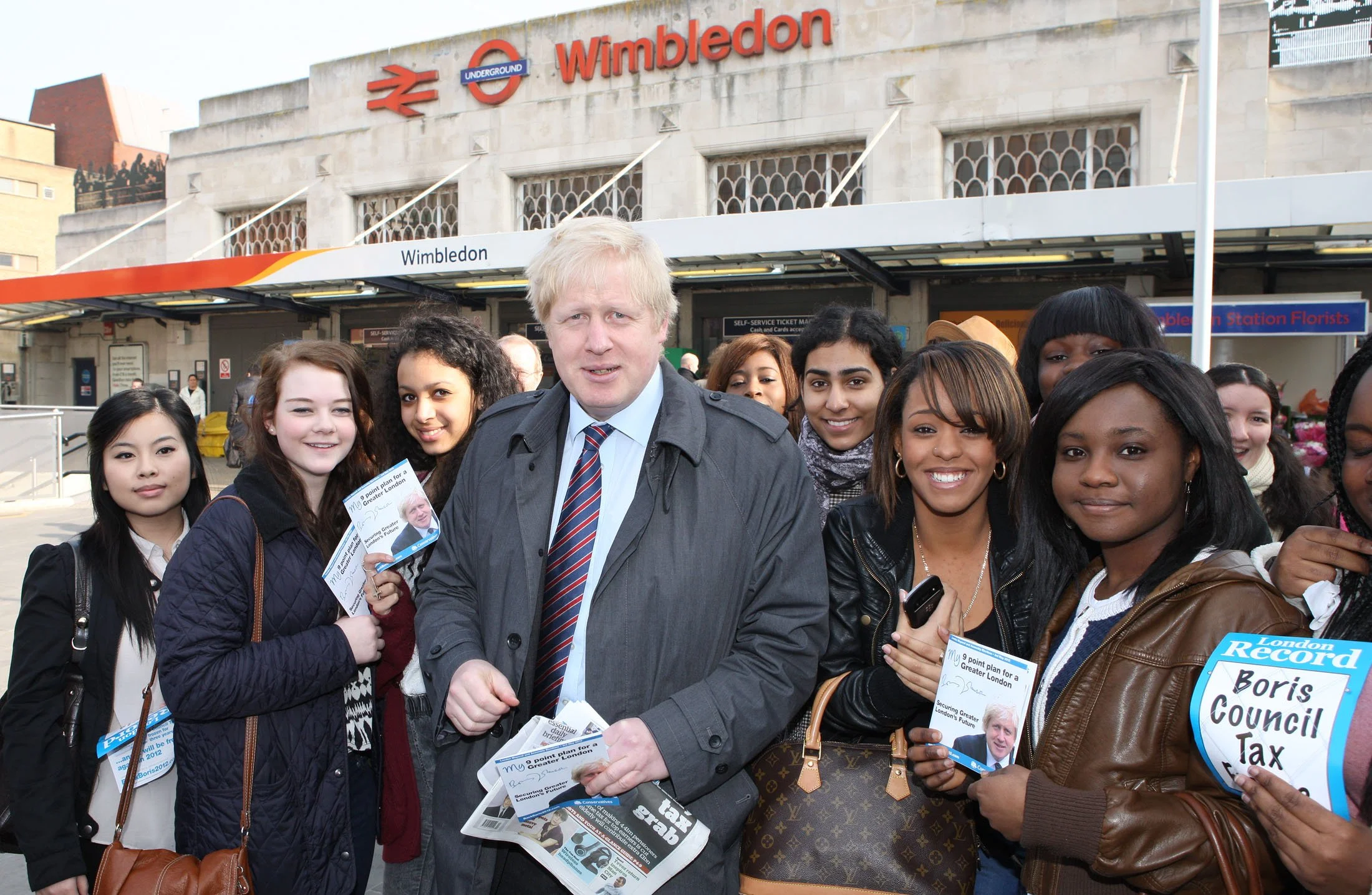 Boris Johnson, the former British Prime Minister, standing with a group of diverse people at Wimbledon station entrance, holding flyers and engaging with the crowd.