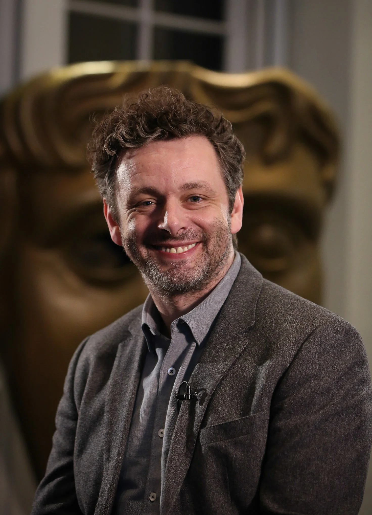 A smiling man with curly hair and a beard wearing a gray shirt and brown blazer.