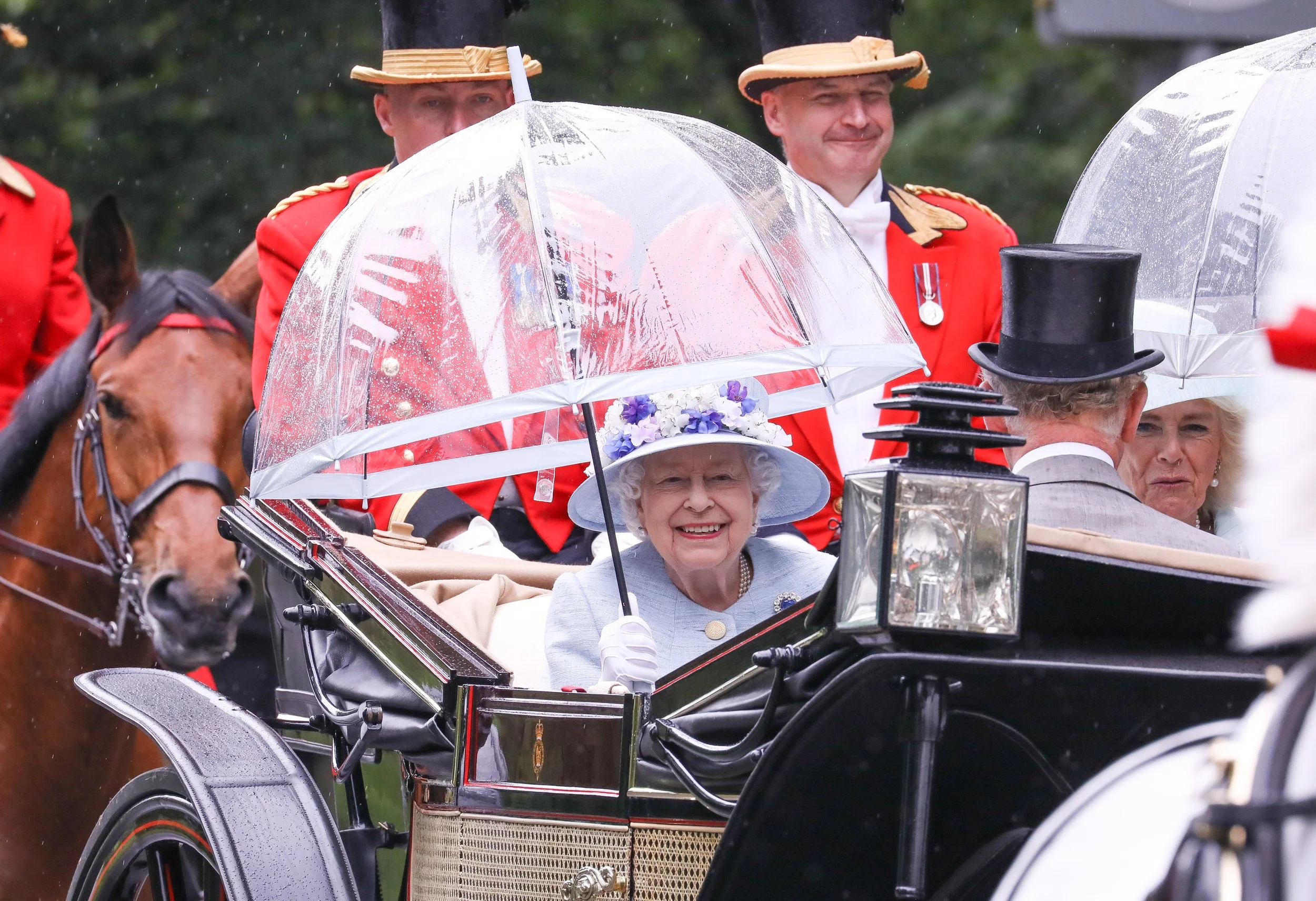 Queen Elizabeth II and others riding in a traditional horse-drawn carriage during a parade, with women holding umbrellas to shield from rain.