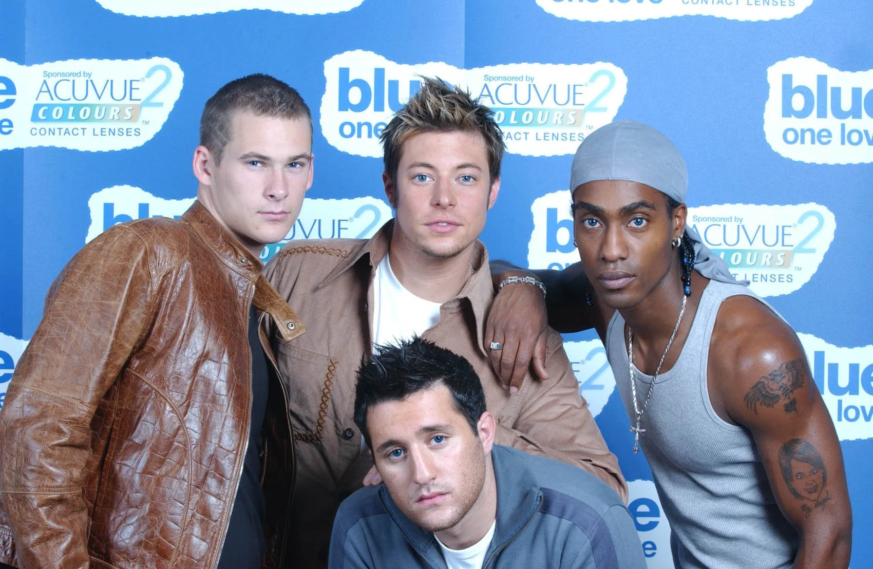 Five men, some with tattoos, posing together against a blue background with 'bluetwo' and 'ACUVUE2 COLORS' logos. One man in a brown leather jacket, another in a brown shirt, another in a gray hoodie, and one in a gray tank top with a gray headscarf.
