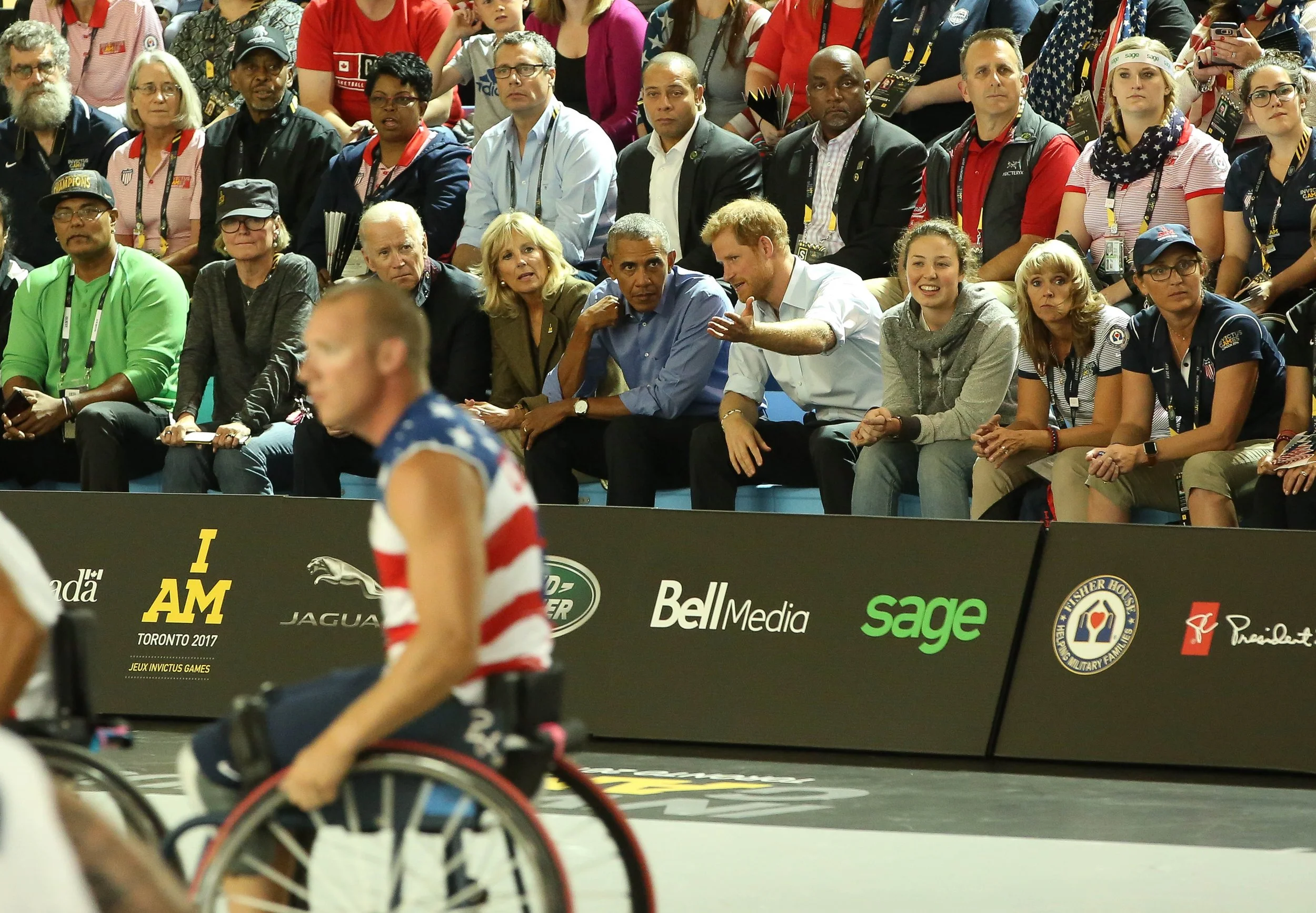 A group of diverse people seated and standing in a sports arena, watching a wheelchair basketball game at the Invictus Games.