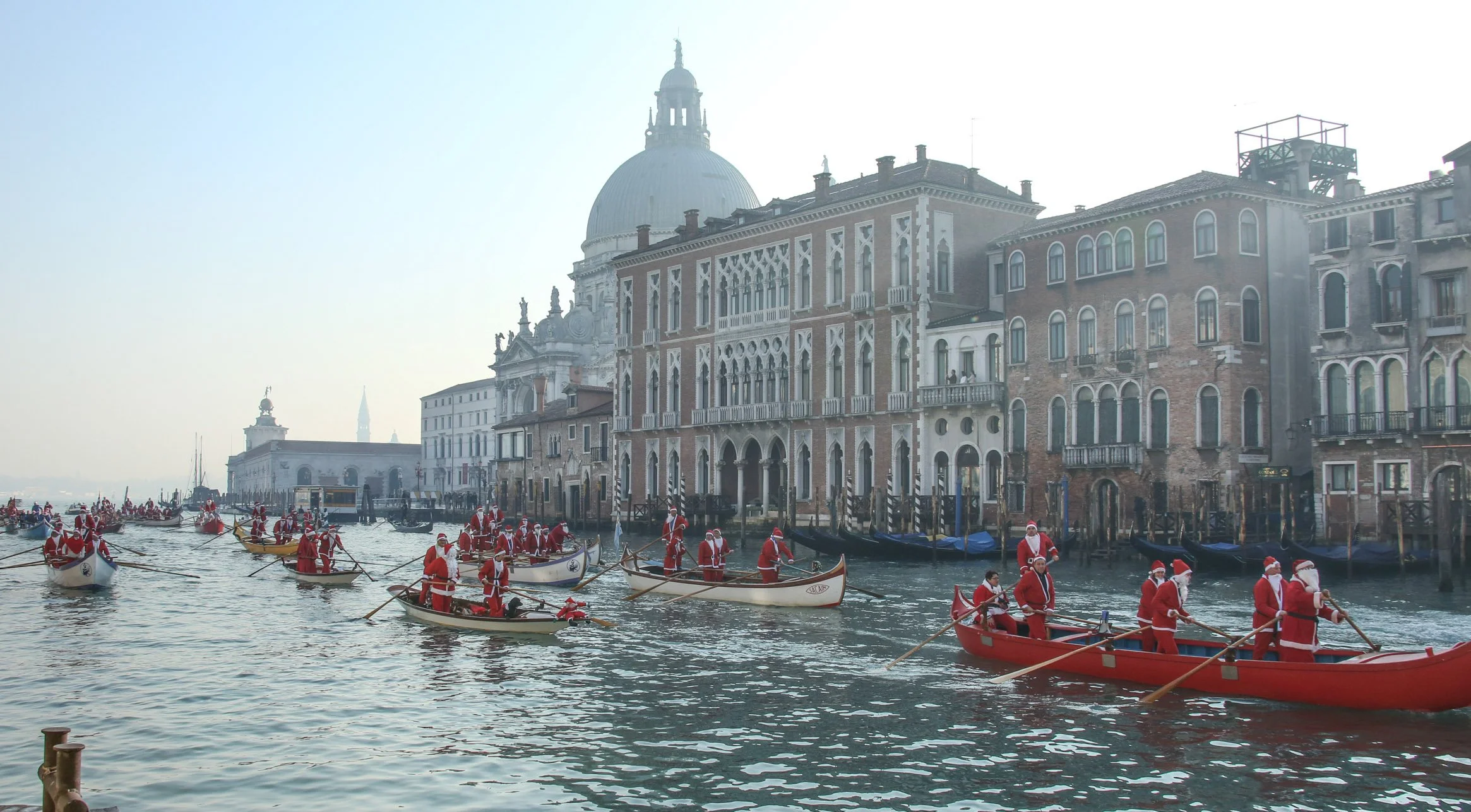 Venetian canal with people dressed as Santa Claus in boats, historic buildings lining the water, and a large domed cathedral in the background.