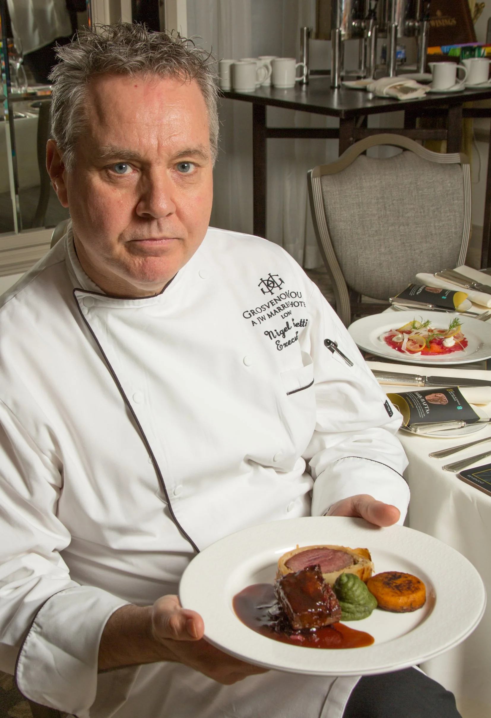 A chef in a white uniform holding a plate with a fancy plated dish, including meat with sauce, vegetables, and garnish in a dining room setting.