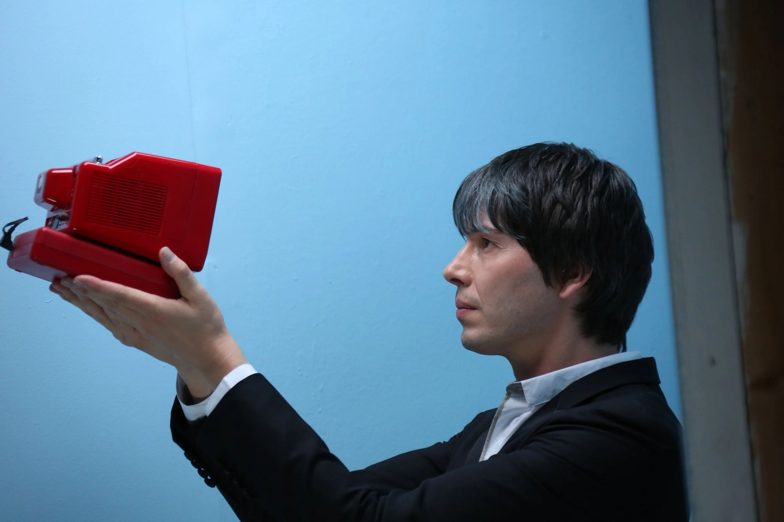 A man in a suit and white shirt holding a red vintage camera, aiming it at the blue wall in front of him.