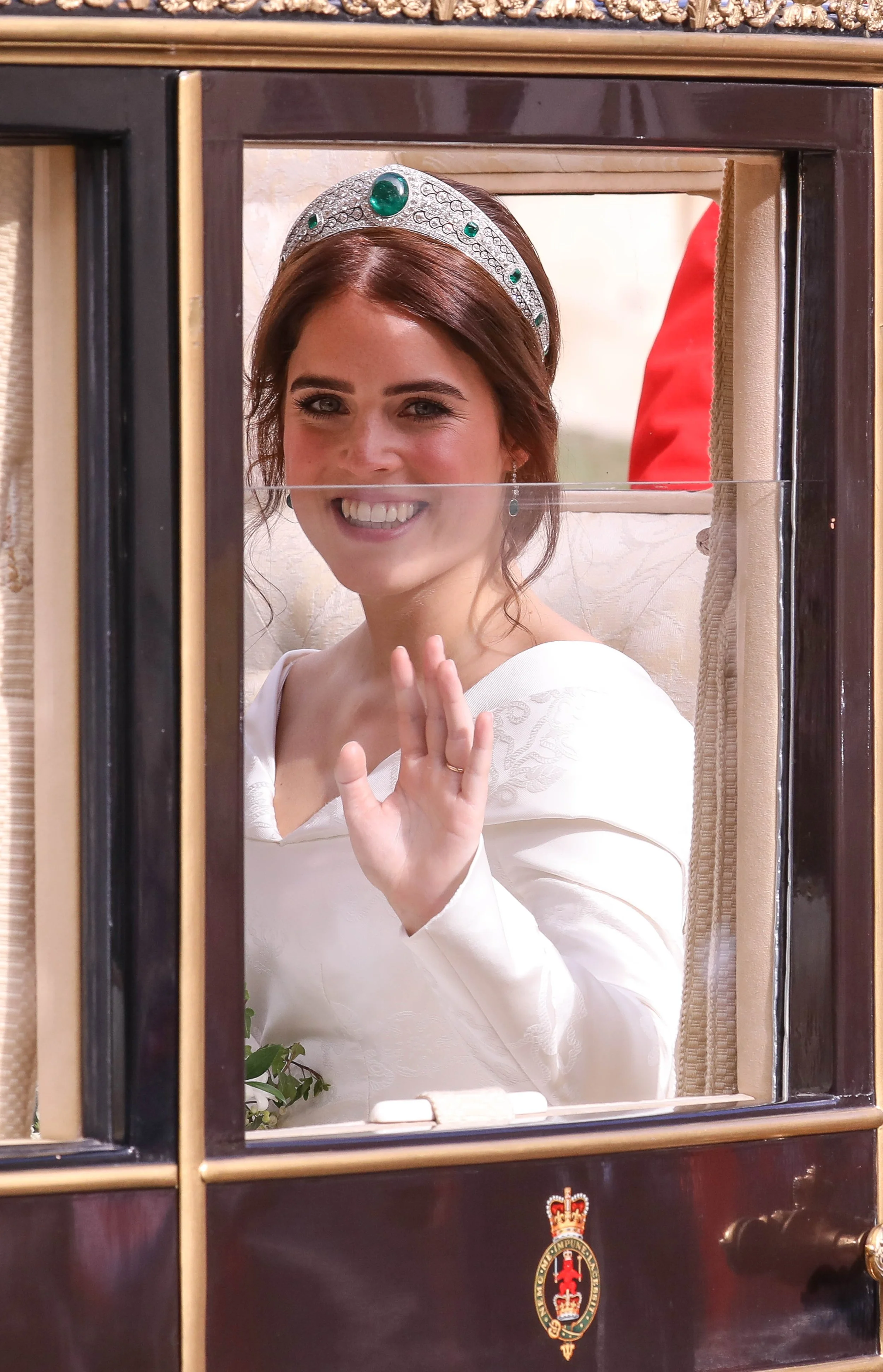 The image shows a woman with brown hair and light makeup, smiling and waving from behind a glass window of a dark wooden carriage with a royal emblem at the bottom. She is wearing a white dress and a tiara with green gems.