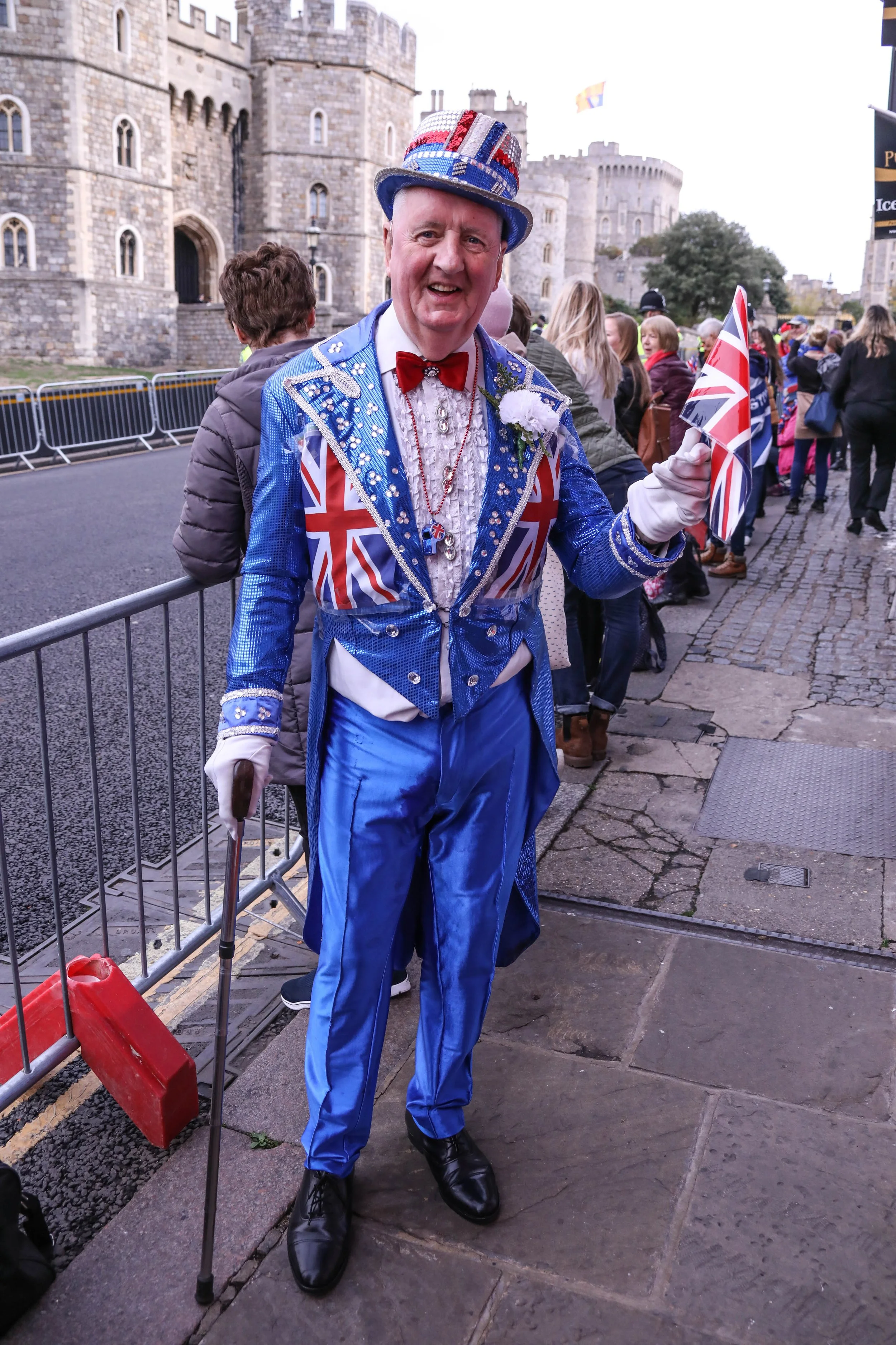 Man dressed in Union Jack themed attire, holding a Union Jack flag, standing outdoors near a crowd with a historic castle in the background.
