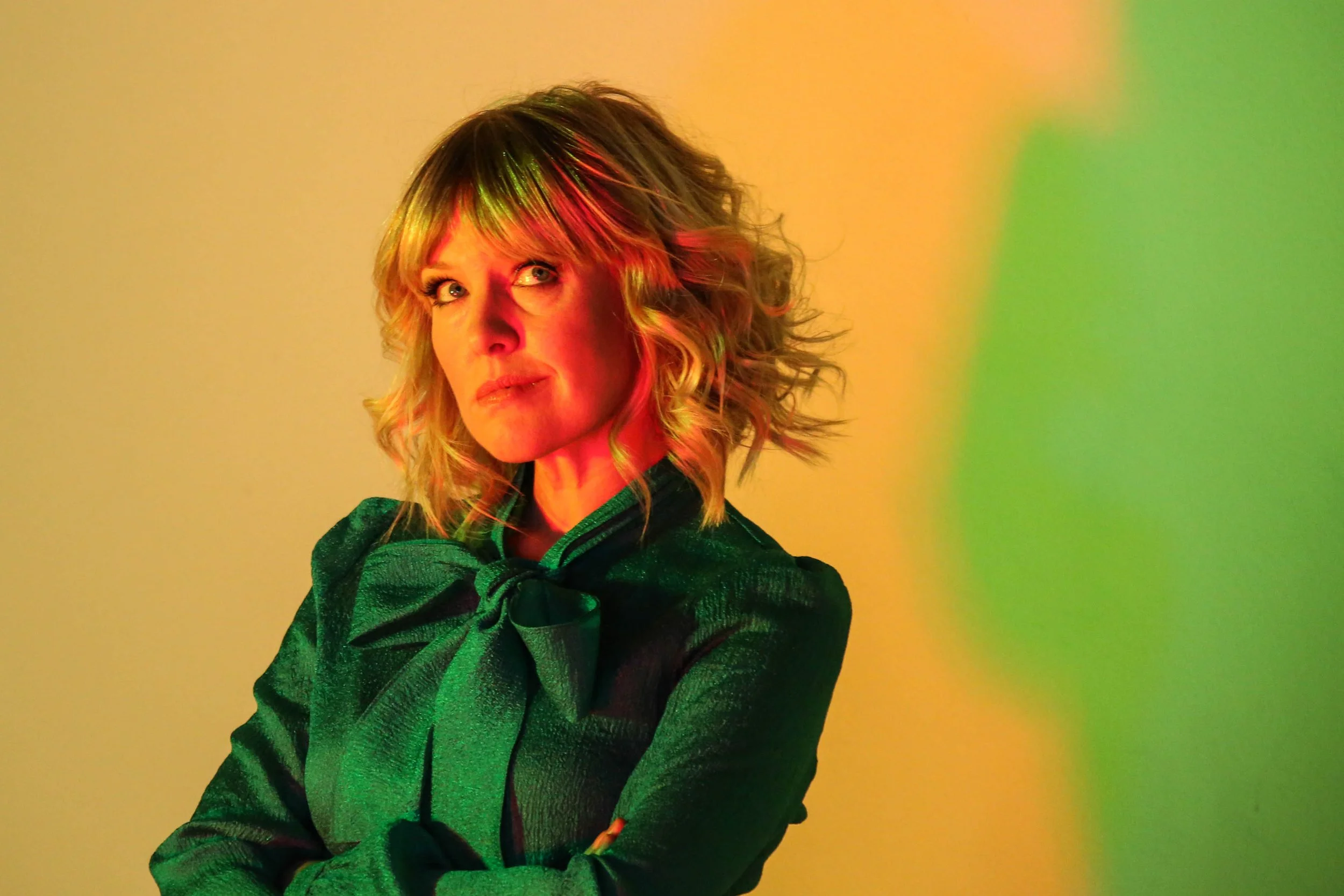 Woman with blonde curly hair wearing a shiny dark green dress with a large bow, standing against a yellow and green gradient background.