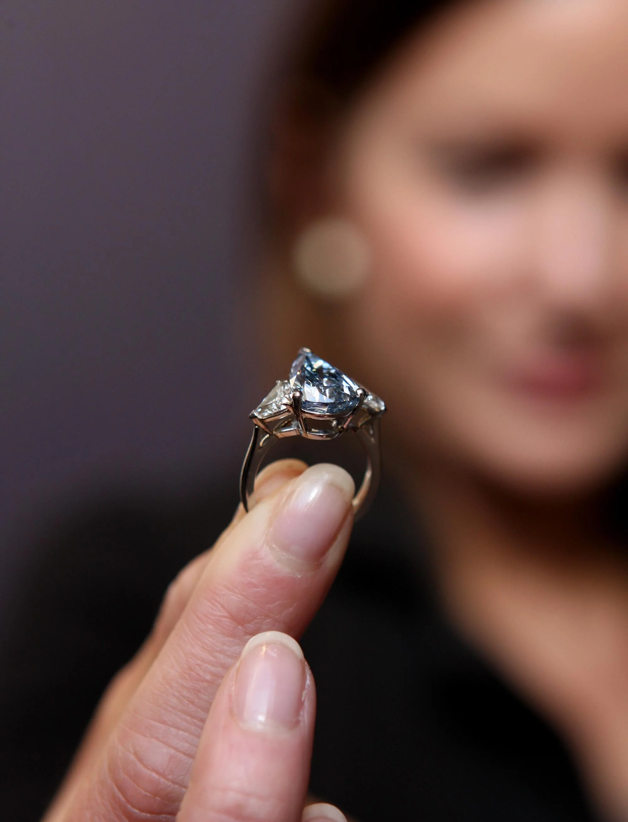 A woman with dark hair and earrings holding a large, faceted diamond engagement ring between her thumb and index finger, with a blurred background.