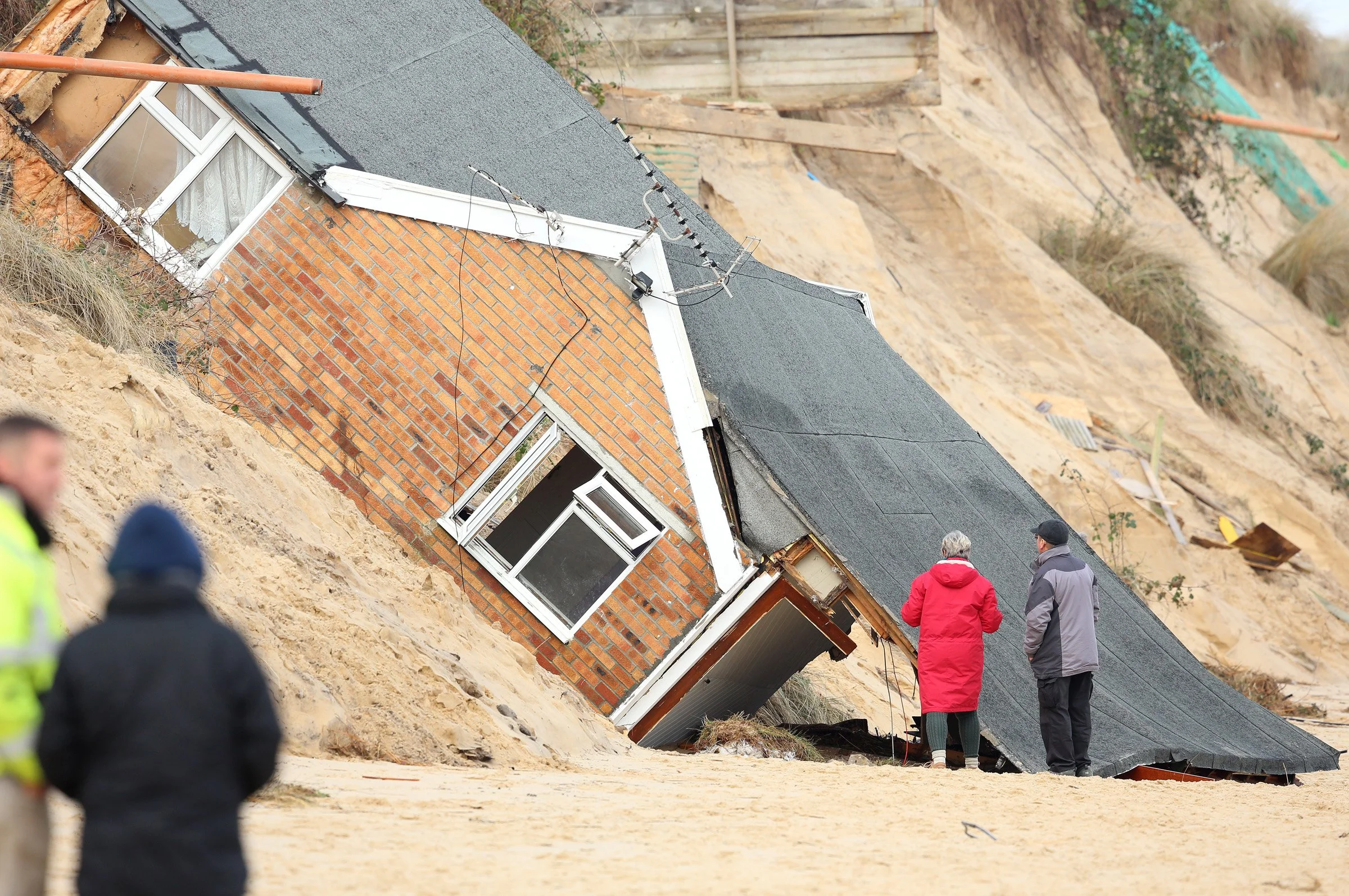 A house with a partially collapsed foundation and tilted walls after land erosion or landslide, with a group of people inspecting the damage outdoors.