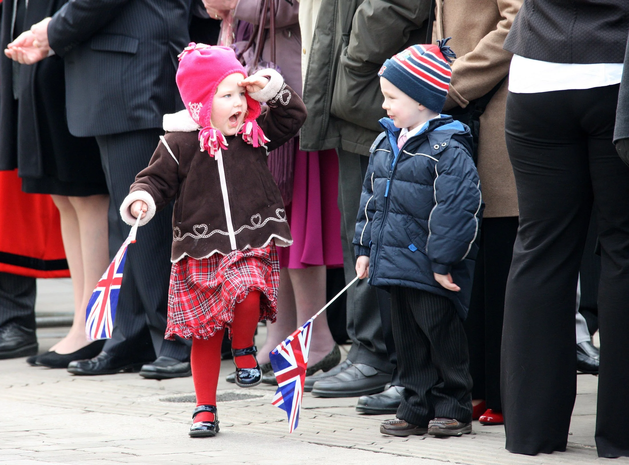 A young girl dressed in festive clothing salutes a parade of people standing in a line, many holding Union Jack flags, during a public celebration or event.