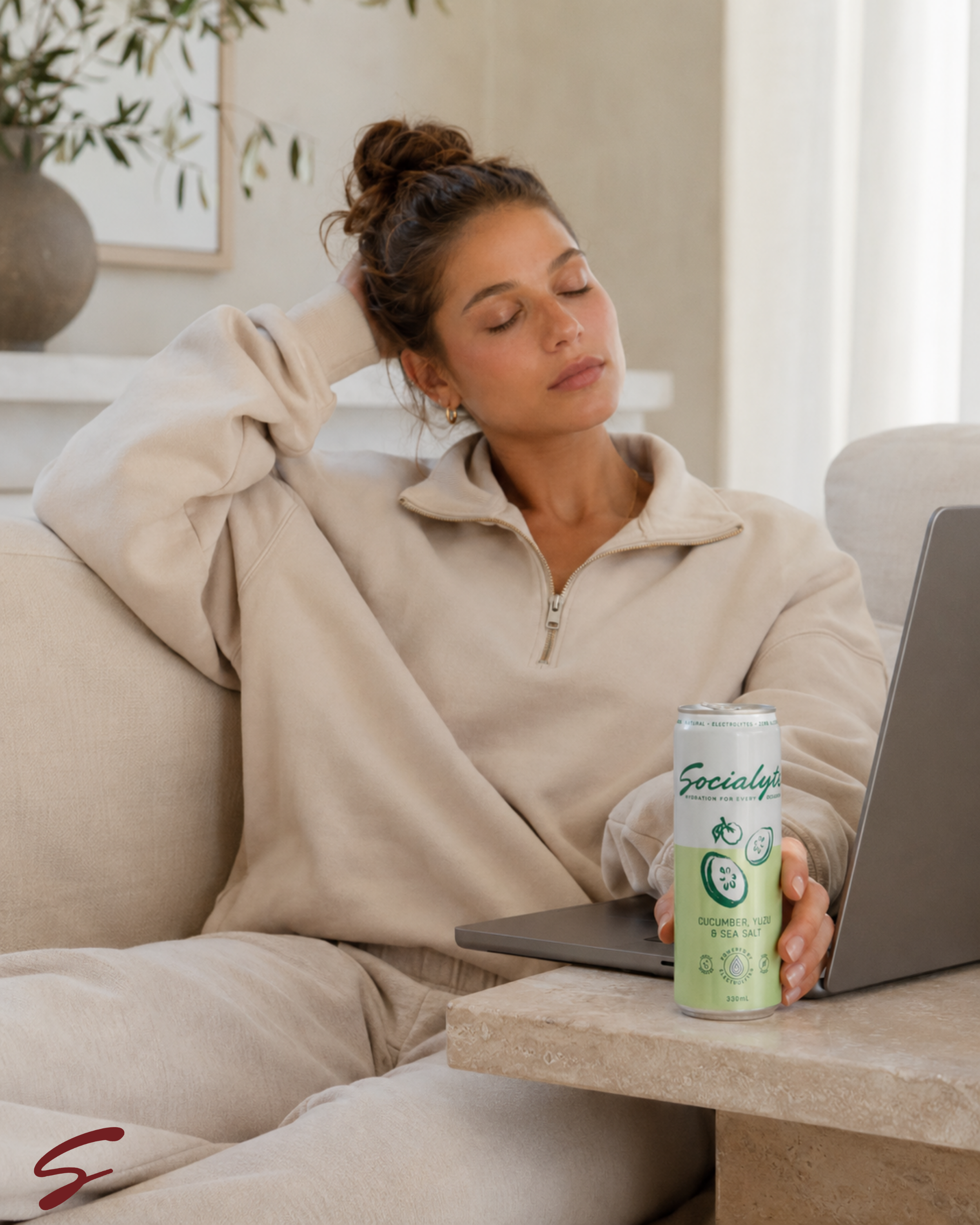 A woman sitting on a beige sofa with her eyes closed, resting her head on her hand. She is wearing a light-colored zip-up hoodie and appears relaxed. On the table in front of her, there is a can of Social Lute beverage with cucumbers, yuzu, and sea s