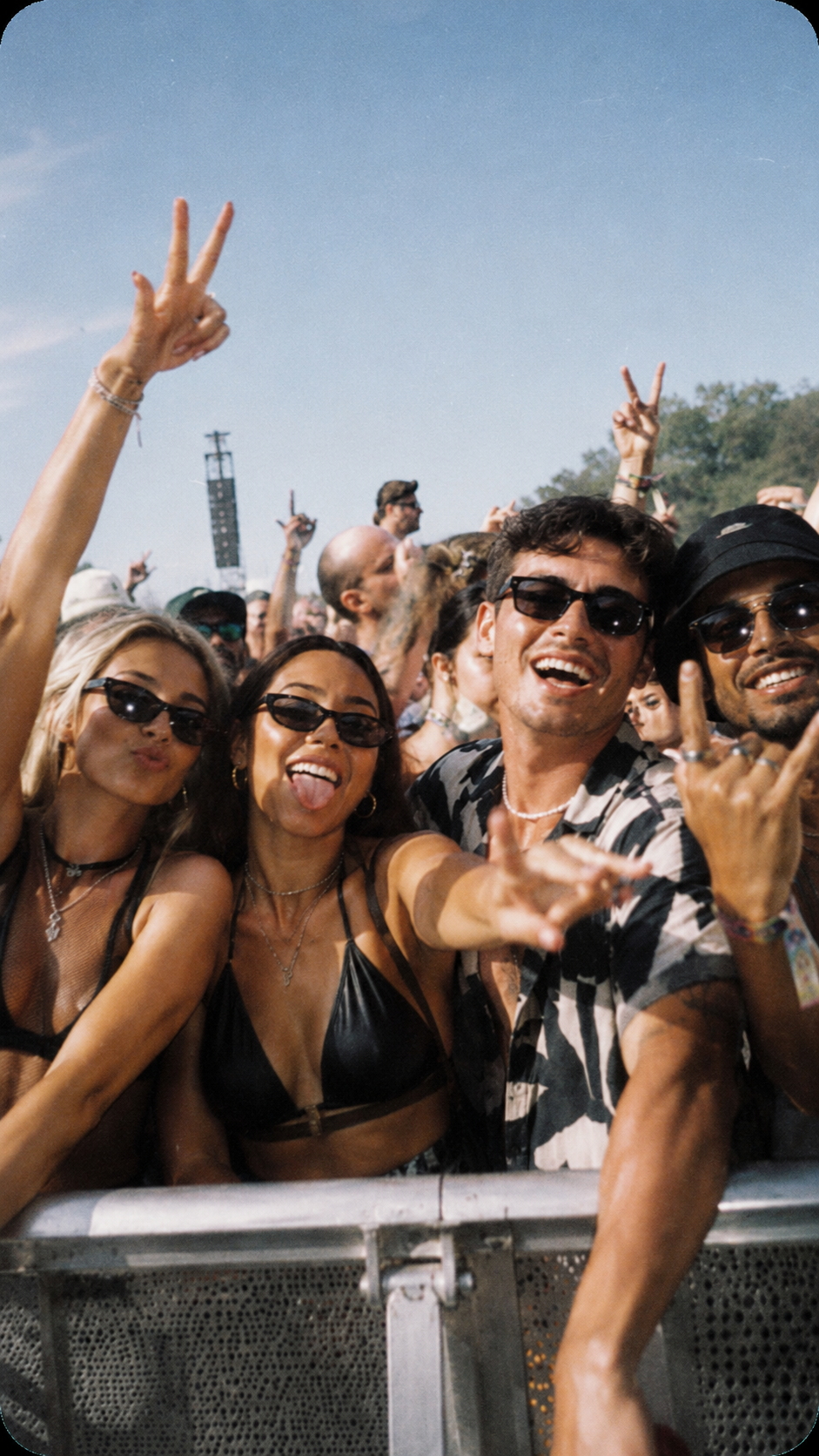 Group of people at an outdoor music festival, smiling and making peace signs, wearing sunglasses and summer clothing, with a crowd and blue sky in the background.