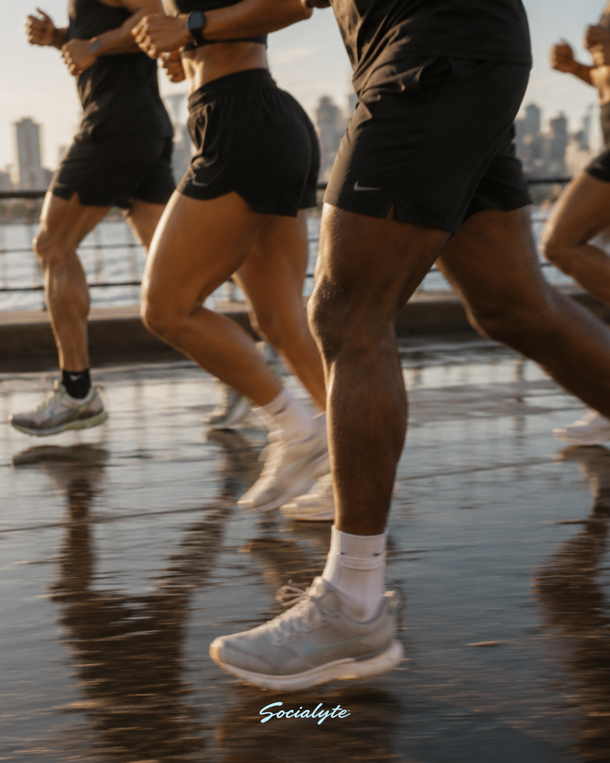 Close-up of multiple runners' lower bodies, wearing shorts and sneakers, running on a wet street in an urban area during sunset.