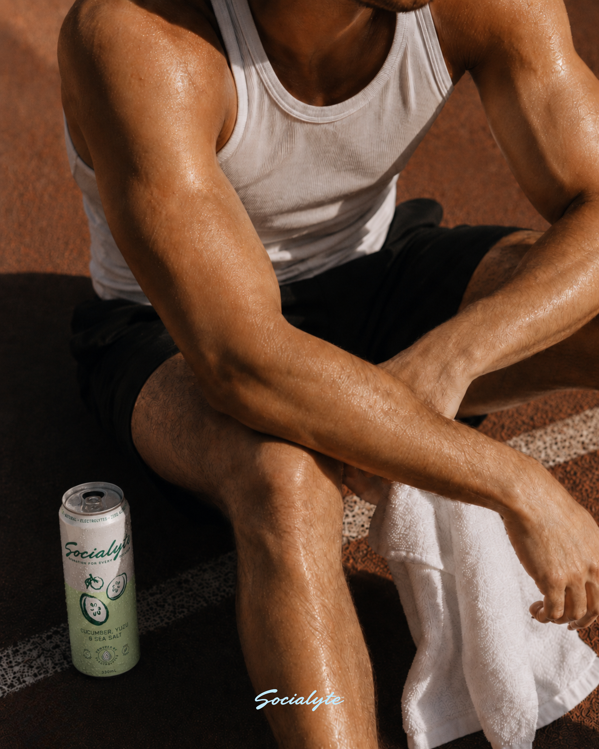 Close-up of man sitting on a brown textured ground, wearing a white tank top and black shorts, with a white towel on the ground beside him. The man's arms and legs are sweaty, and he appears to be in a resting or post-exercise position. There is a wh