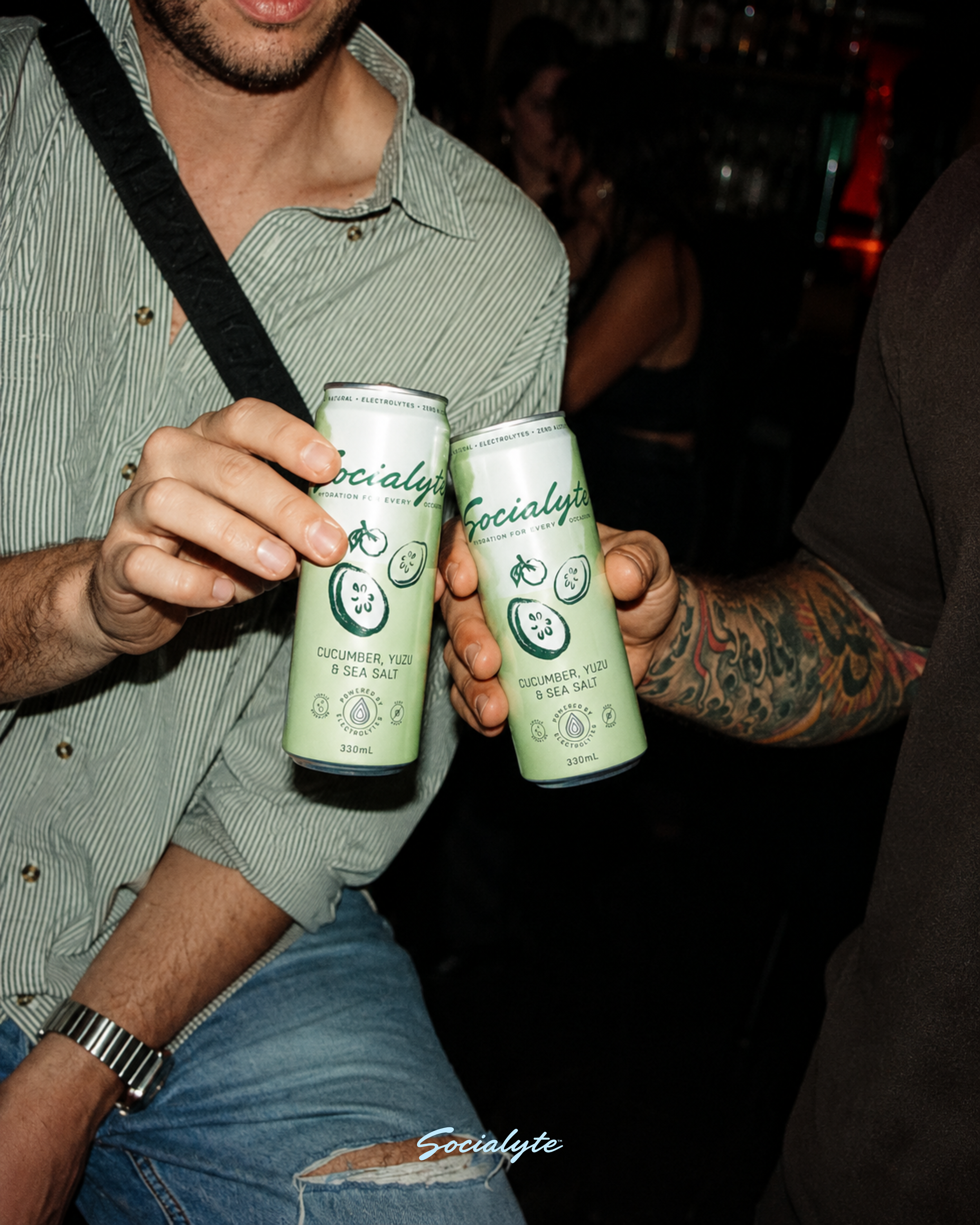 Two people holding cans of Socialite cucumber, yuzu, and sea salt sparkling water at a social event or bar. One person is wearing a green and white striped shirt, the other has a tattooed arm visible. The background is dark with blurred figures.