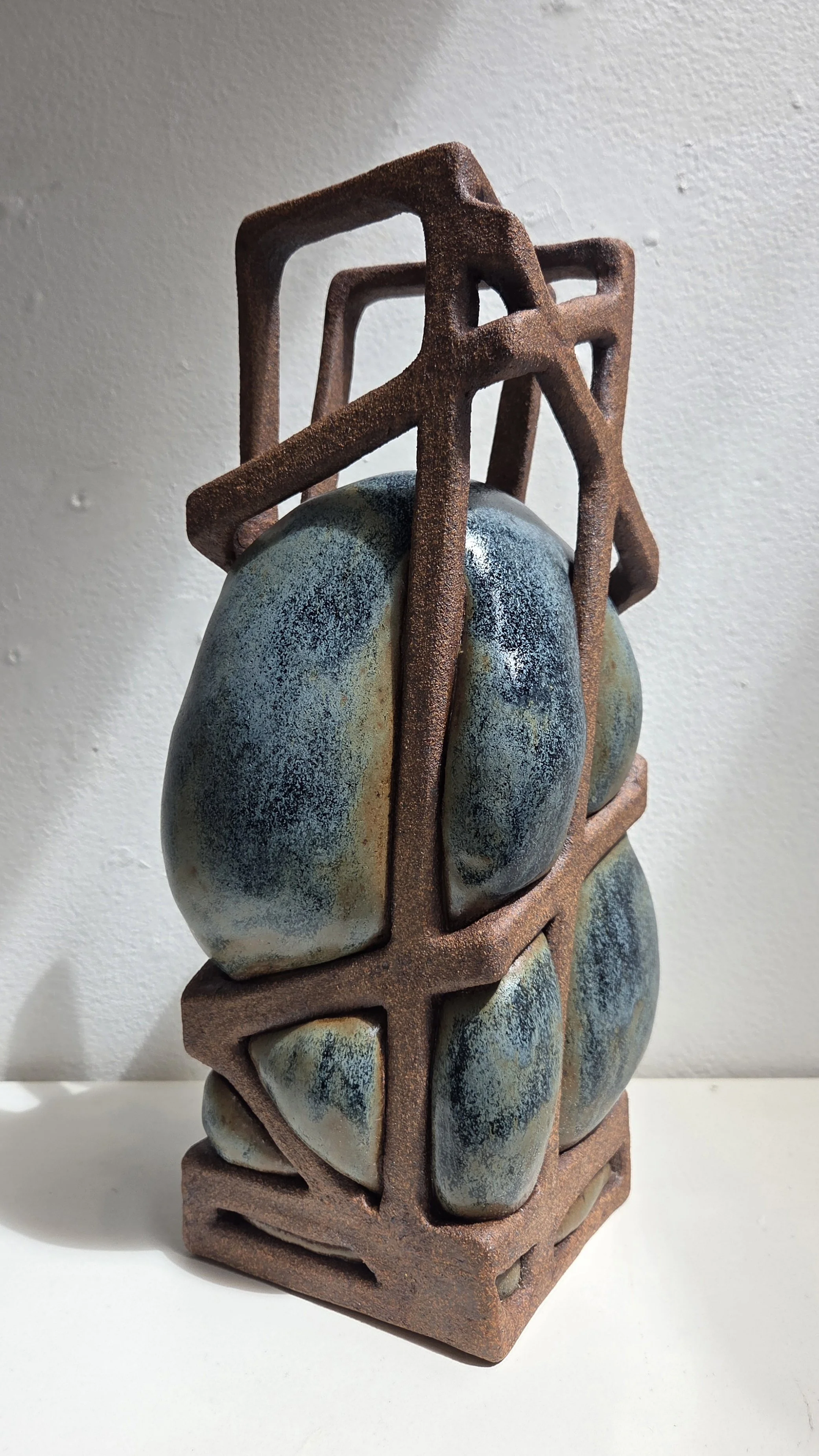 Sculpture featuring smooth, blue green glaze buldges held within a rusted ceramic frame that looks rusty.