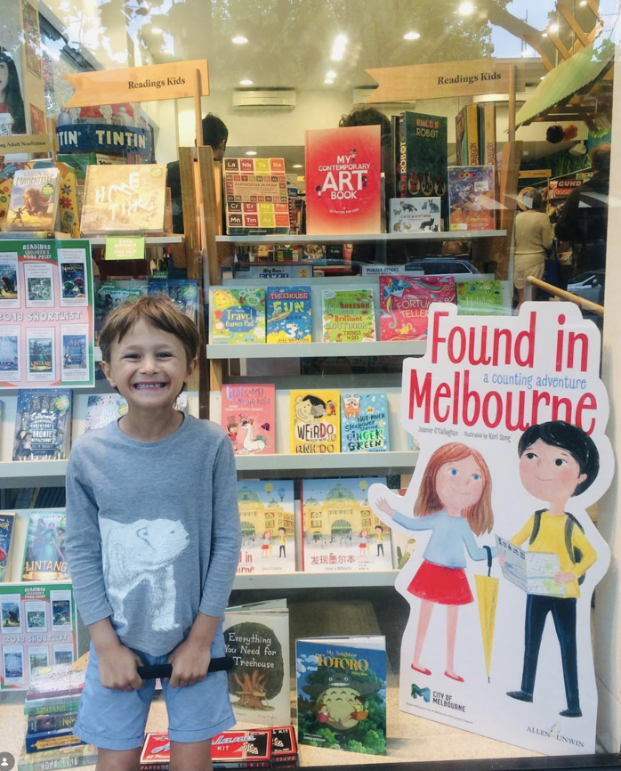 Young boy smiling standing in front of a bookstore display for children's books, with a sign that says 'Found in Melbourne' featuring illustrations of two children, one girl and one boy.