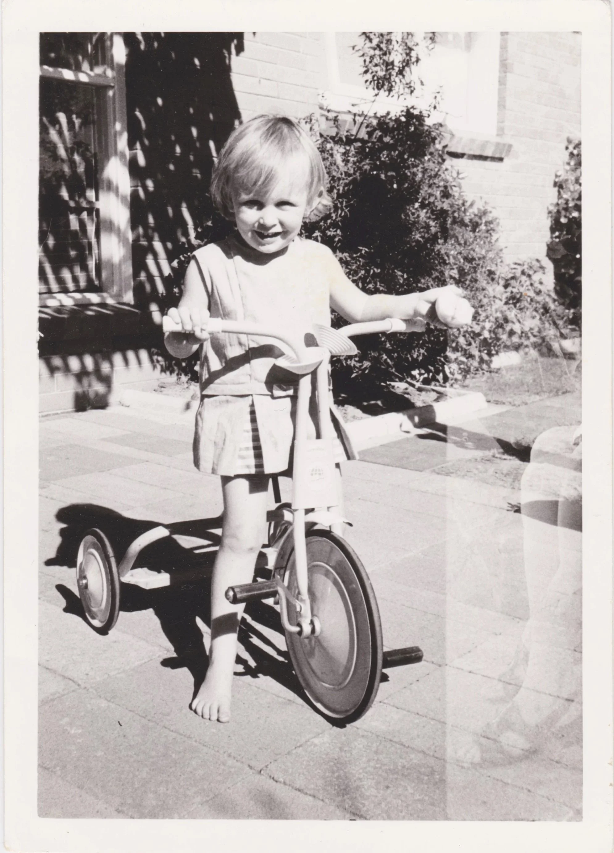 A young girl with blonde hair riding a tricycle outdoors in front of a house with brick walls and bushes. She is smiling and appears to be enjoying herself.