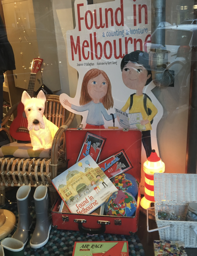 Display window with a children's book titled "Found in Melbourne" and various travel-themed items, including a plush dog, a red guitar, rain boots, a toy lighthouse, and a small globe. Illustration of a girl and boy with a backpack on the poster.