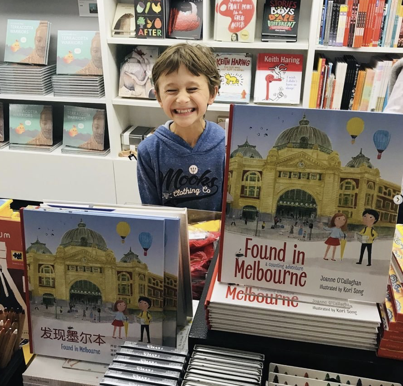 A young boy with a big smile, wearing a blue hoodie, standing behind a table with copies of the book titled 'Found in Melbourne'. The book cover shows a historic building, two children, and hot air balloons. Bookshelves with various books are in the 