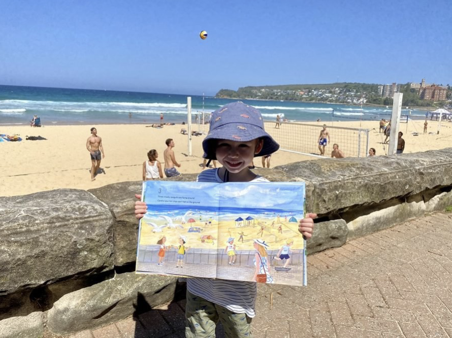 Child wearing a bucket hat smiling and holding an illustrated book at the beach, with people playing volleyball and relaxing in the background.