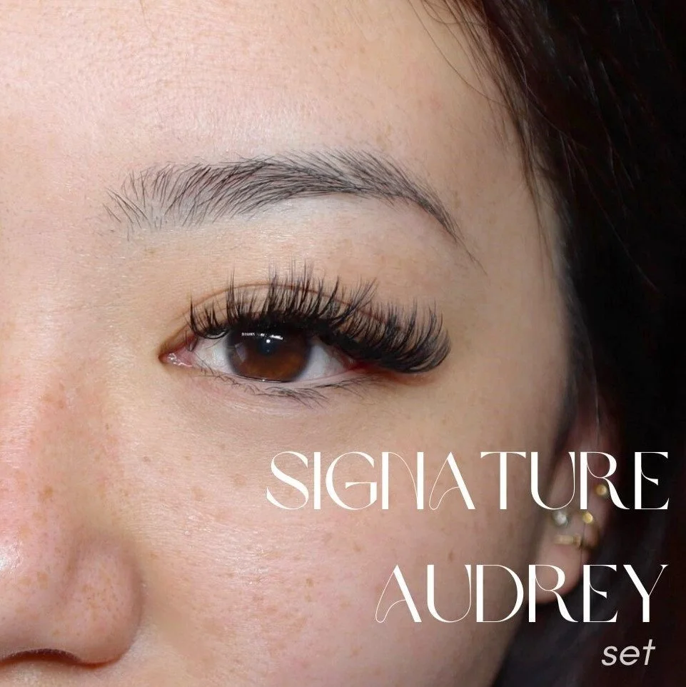 Close-up of a woman's face focusing on her eye, showcasing long eyelash extensions. The image includes the text 'SIGNATURE AUDREY set' in white letters.