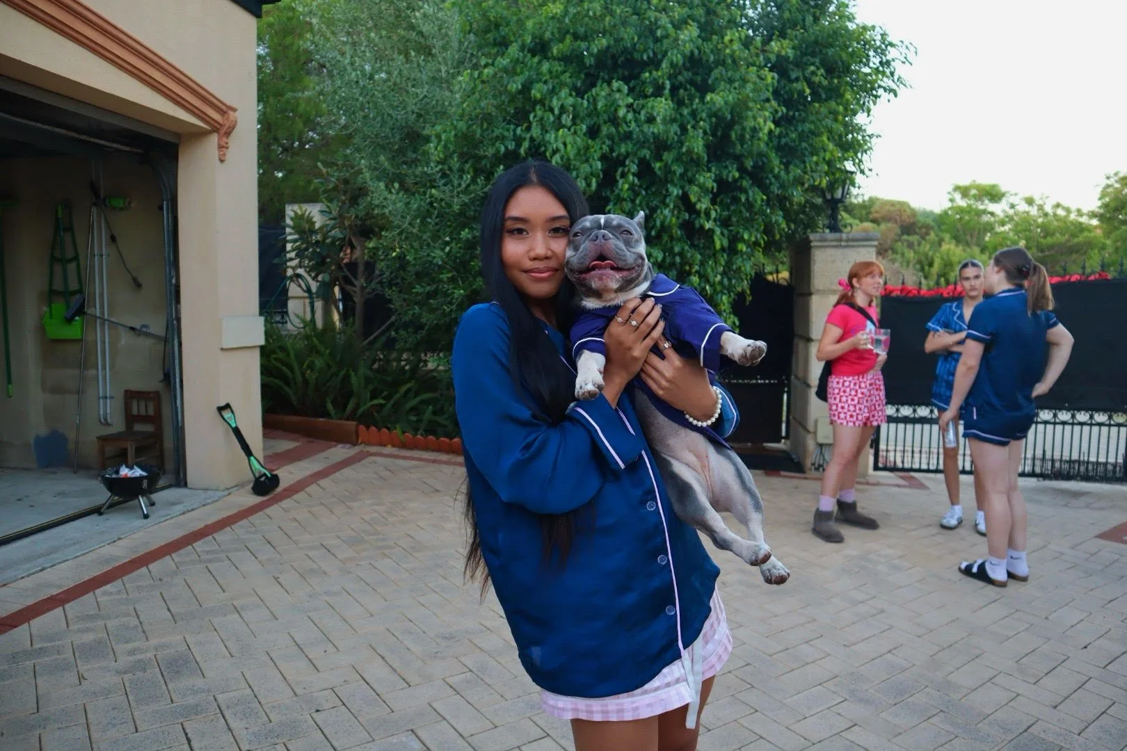 A woman holding a smiling dog in a backyard with three girls standing in the background, green trees, and a black fence.