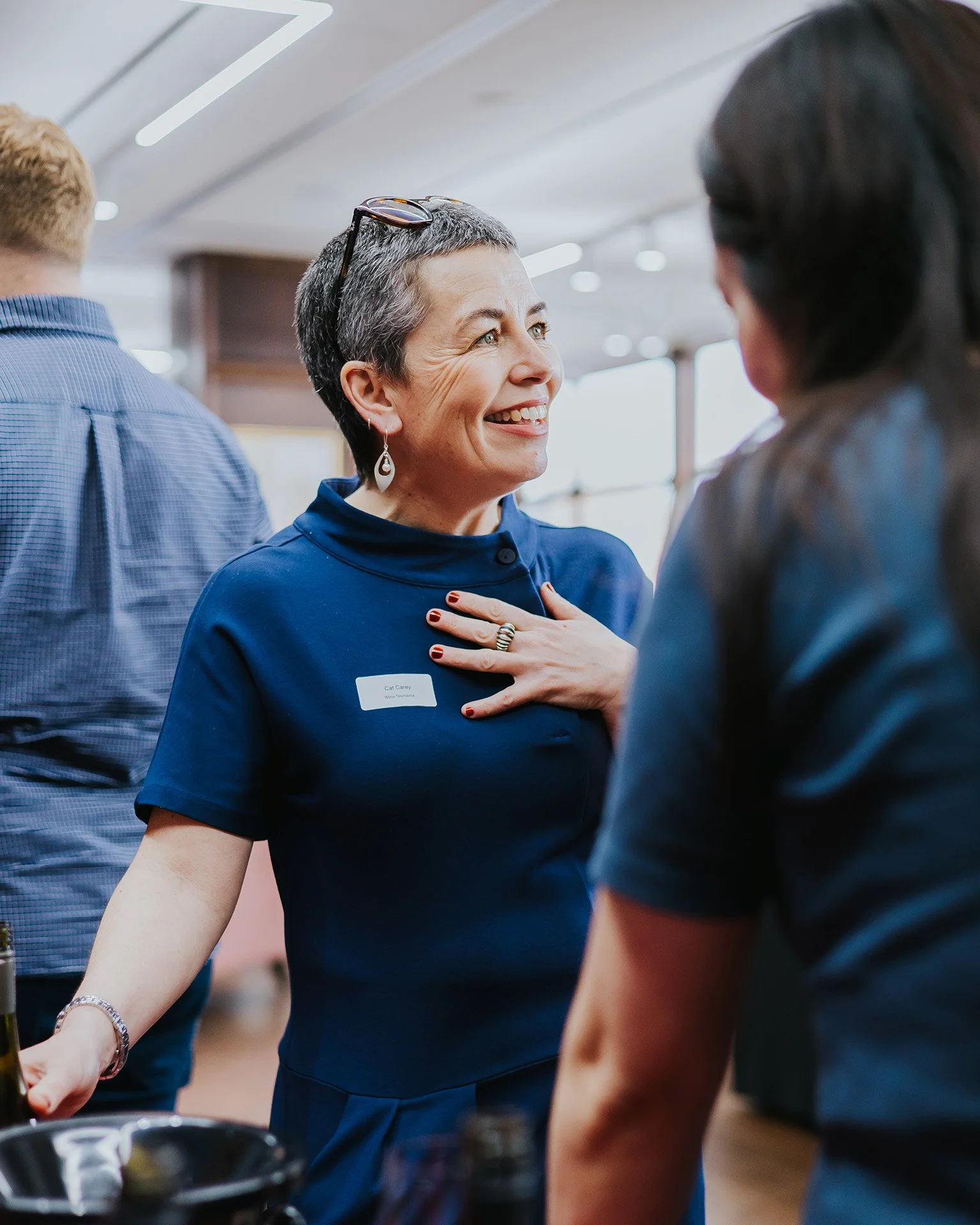 A woman with short gray hair, wearing a dark blue shirt with a name tag, smiling and engaged in conversation with a person whose back is turned.