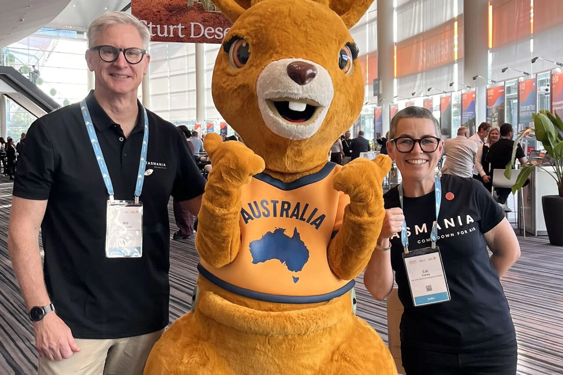 Two people and a person in a kangaroo costume posing at an indoor event. The man is on the left, wearing glasses, a black polo shirt, and beige pants. The woman is on the right, wearing glasses, a dark shirt, and a lanyard. The kangaroo costume has a yellow-orange body, large eyes, and a shirt with an outline of Australia. The background shows large windows and a busy environment with other attendees.