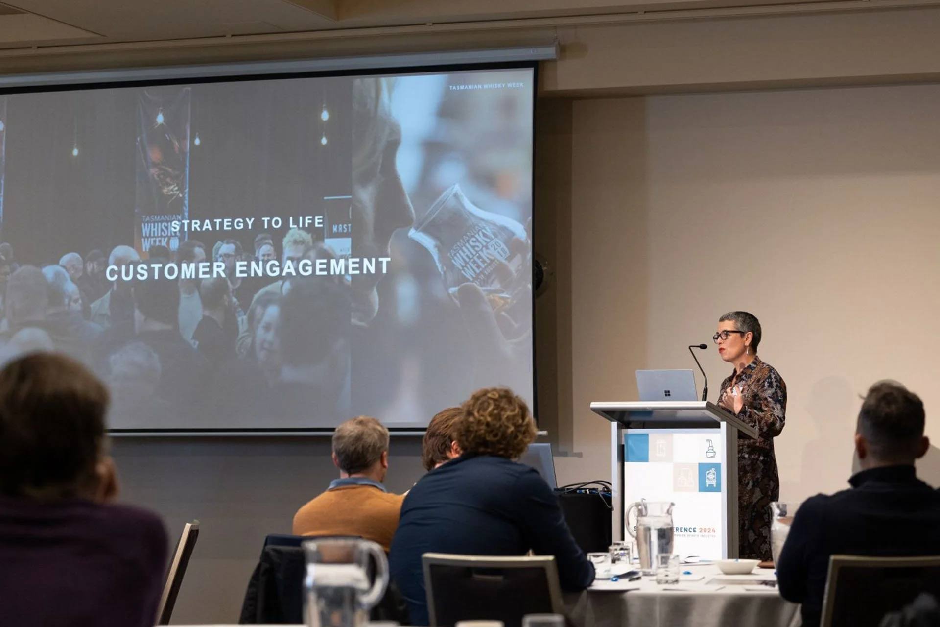 A woman with short gray hair and glasses standing at a podium giving a presentation in a conference room. A large screen behind her displays a slide that reads 'Strategy to Life Customer Engagement' with an image of people at a whisky event. Audience members are seated and facing the speaker.