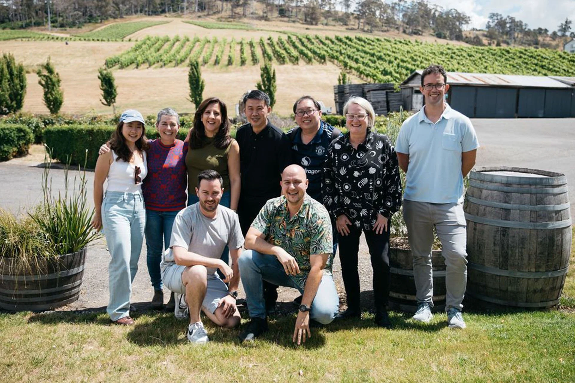 Group of ten people posing outdoors in front of a vineyard on a sunny day. Three are seated on the grass, while the others stand behind. Tall barrels and a small wooden shed are visible nearby.
