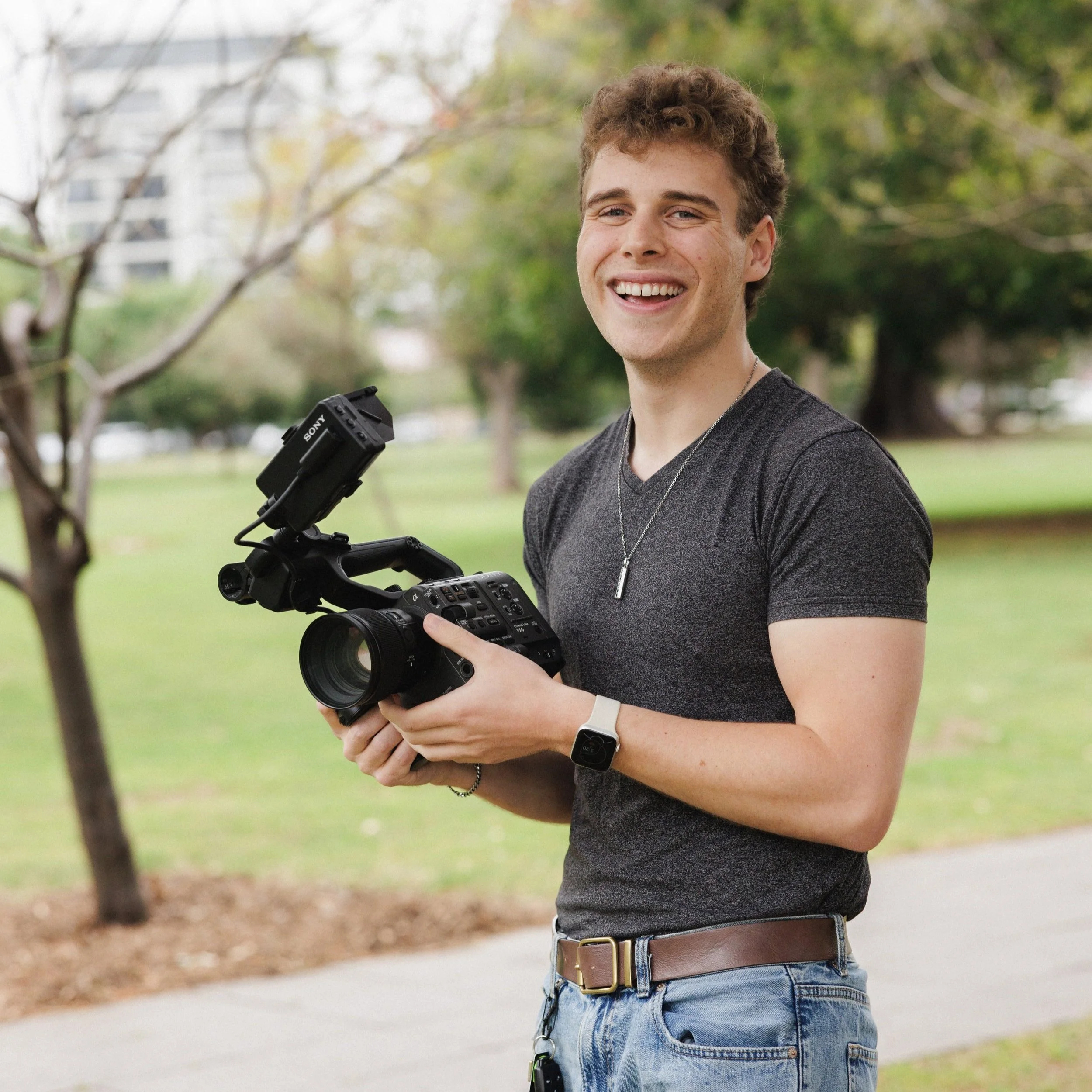 Person outdoors holding a video camera, smiling, wearing a gray T-shirt and blue jeans, with trees and grass in the background.
