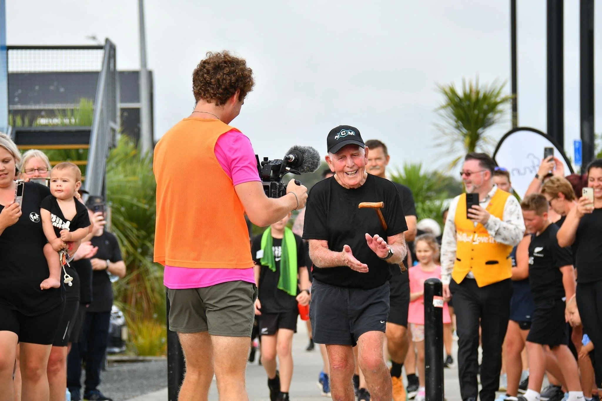 Elderly man in running attire smiling, surrounded by a crowd, while a person films him with a camera. People in the background clapping and taking photos.