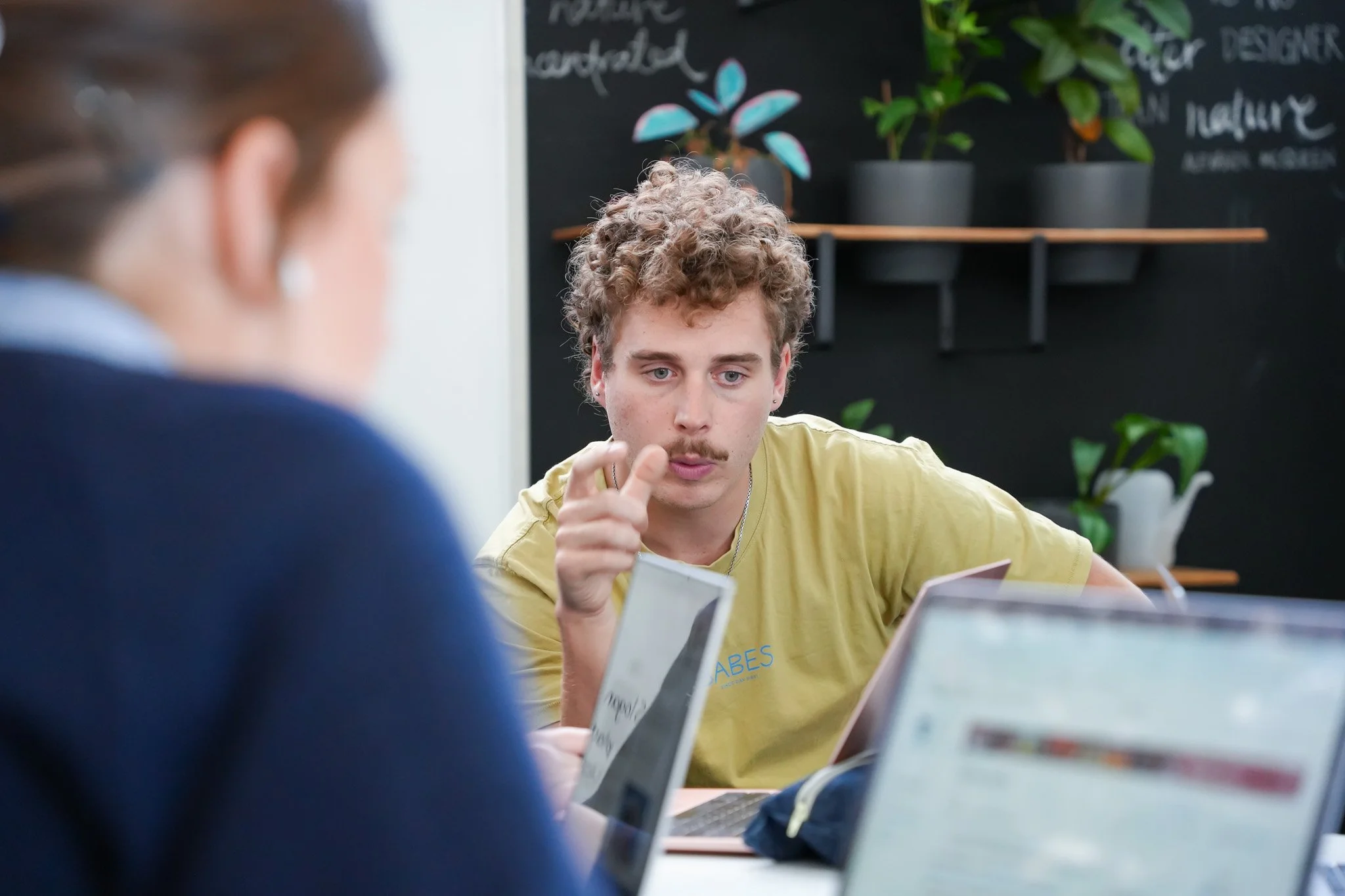 A man with curly hair and a mustache is sitting at a table, gesturing with his hand, surrounded by laptops and plants on shelves in the background. Another person is partially visible in the foreground.