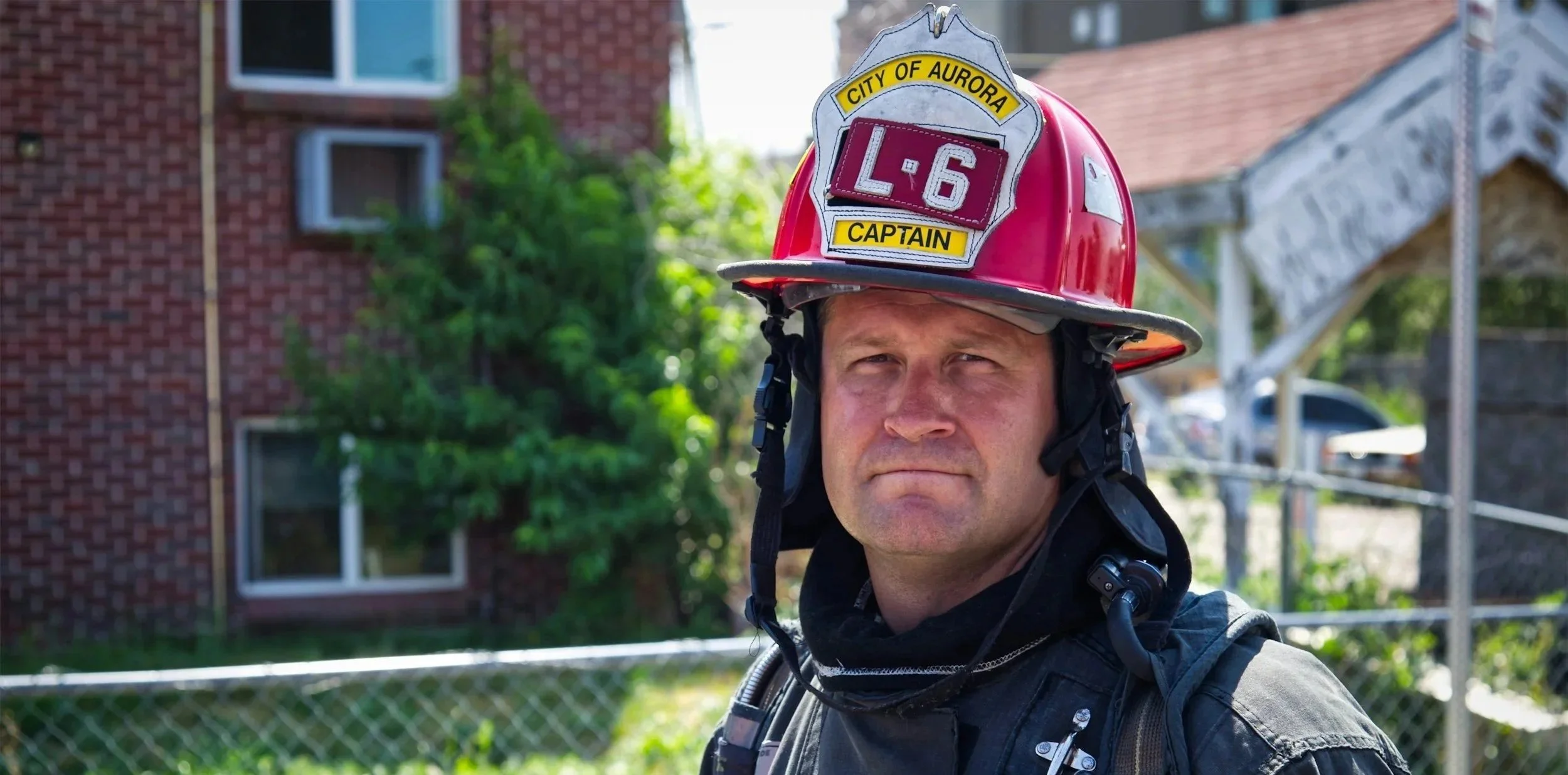 Aurora, Colorado, firefighter with a red helmet labeled 'L-6' and 'Captain' stands outdoors in front of a brick building and trees.