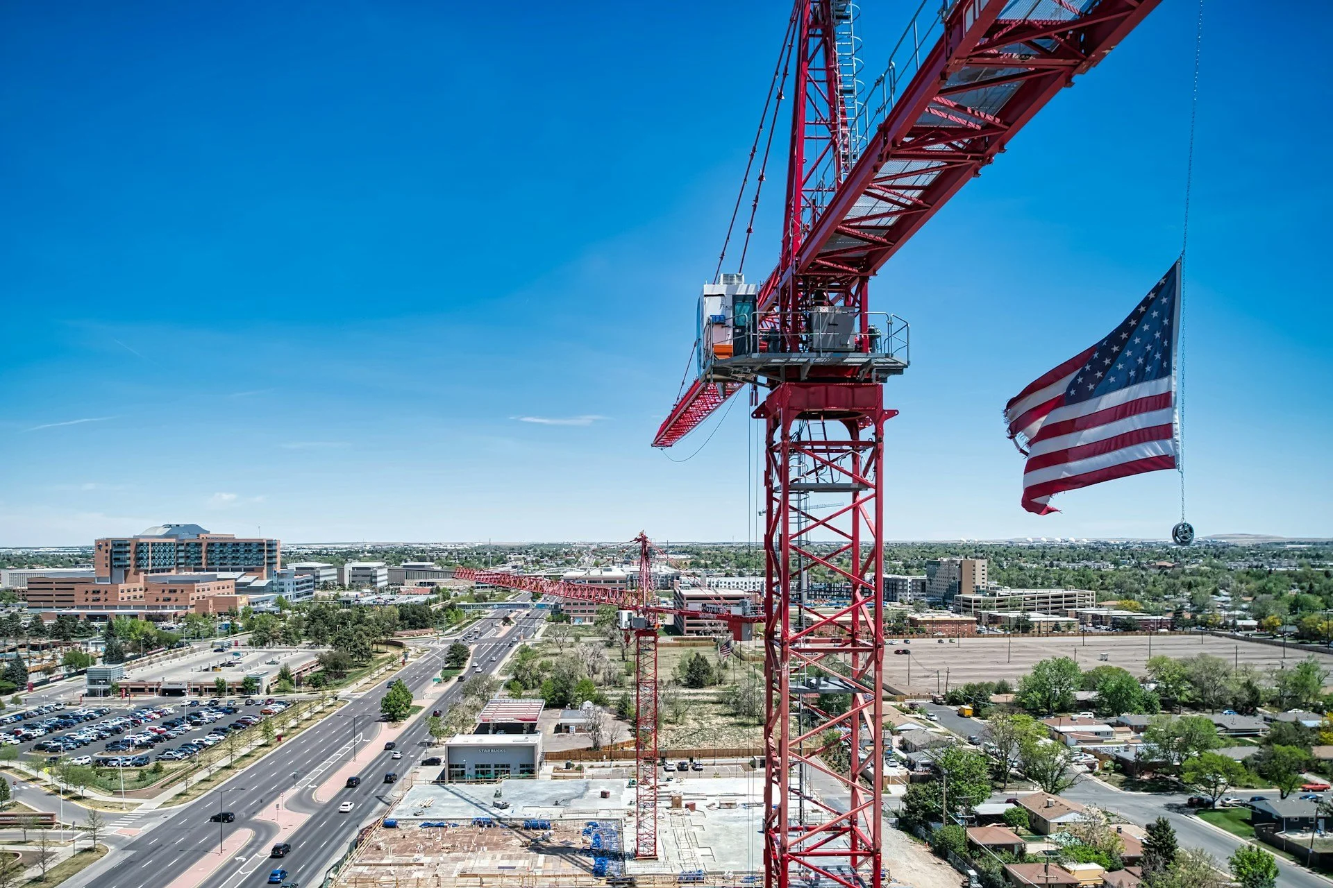 Construction crane with American flag flying in clear blue sky over Aurora, Colorado, cityscape and roads.