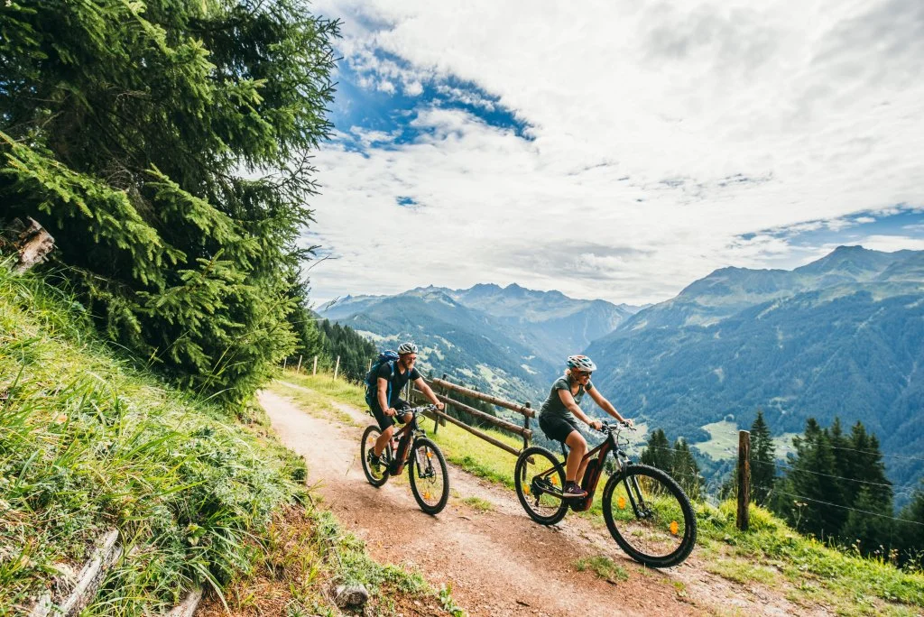 Zwei Mountainbiker fahren auf einem Trails in den Bergen, umgeben von Wäldern und Blick auf Berge und Wolken am Himmel.