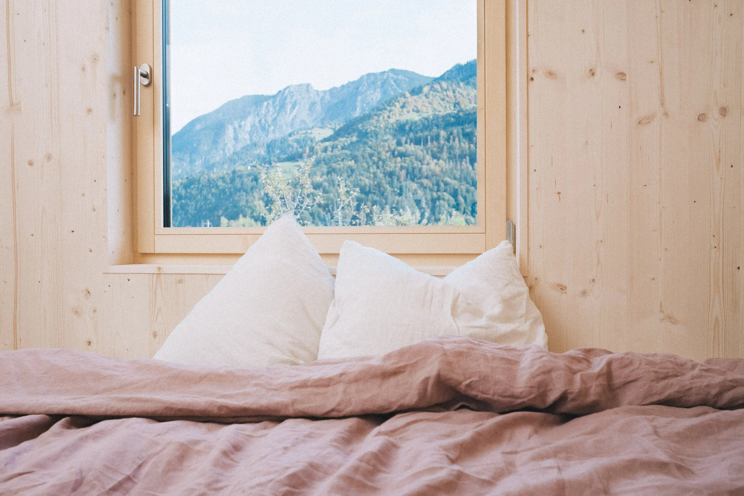 Blick aus einem Fenster auf Hügel und Berge in der Ferne, mit weißen Kissen auf einem Bett im Vordergrund, in einem Holzzimmer.