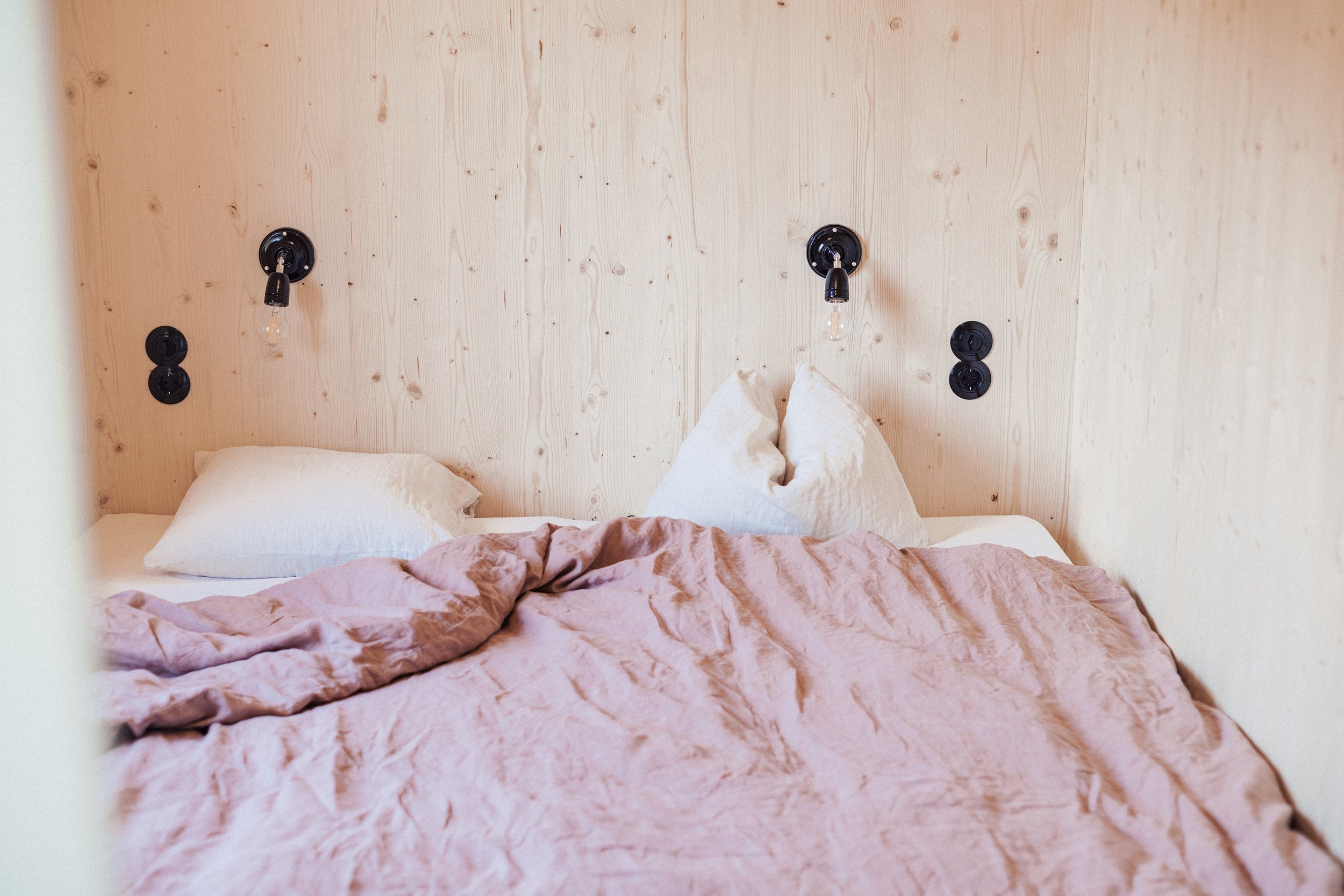 Bett mit weißen Kissen und verzerrtes rosa Bettlaken vor einer Holzwand mit Wandlampen