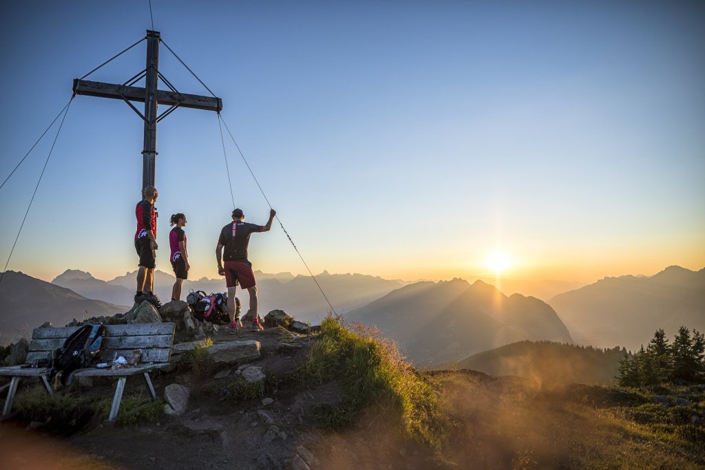 Drei Personen stehen auf einem Berggipfel bei Sonnenuntergang, vor einem großen Holzkreuz, mit Rucksack und Trekkingausrüstung, mit Blick auf die Berglandschaft.