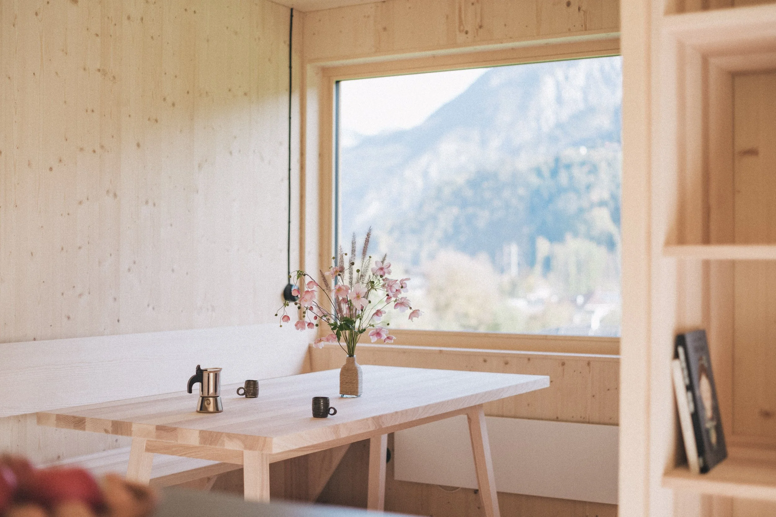 Ein Holzarbeitsplatz mit einer Vase voller pinker Blumen, eine kleine Kanne und eine Thermoskanne vor einem großen Fenster mit Bergblick in einem Holzhaus.