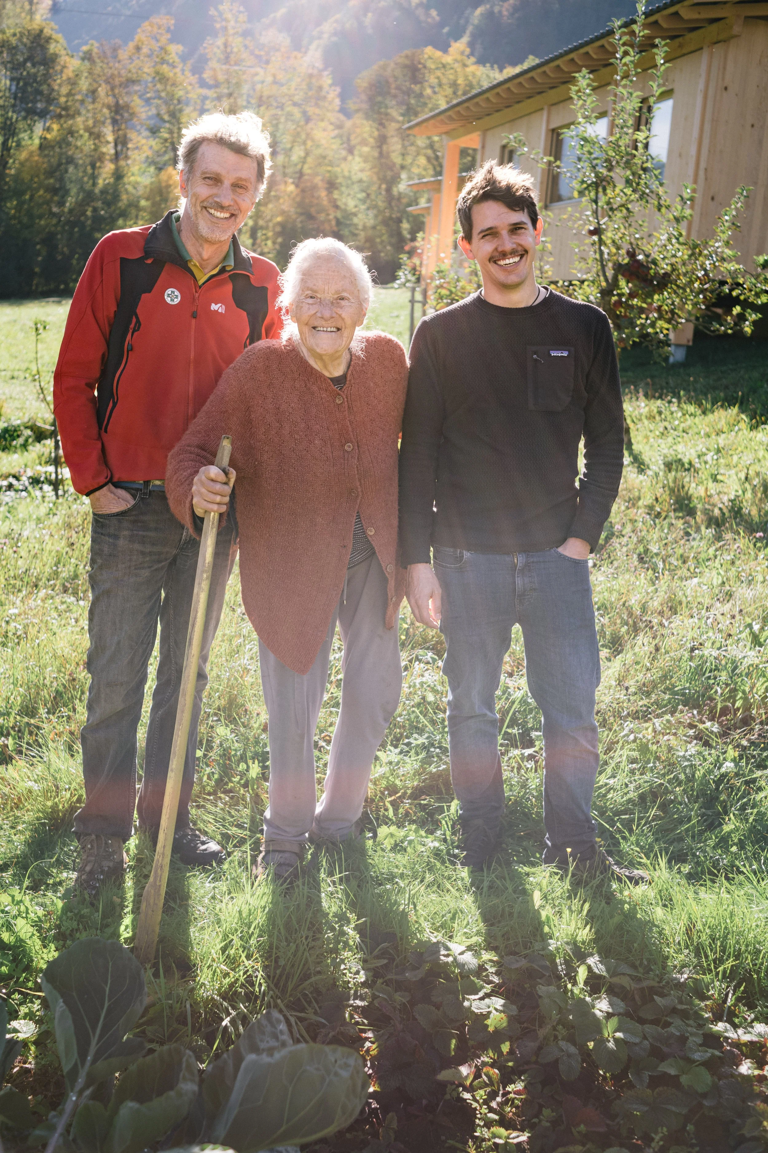 Vier Menschen, zwei Männer und eine ältere Frau, stehen im Garten vor einer Holzhütte und lächeln. Es ist sonnig und die Bäume im Hintergrund haben Herbstfarben.