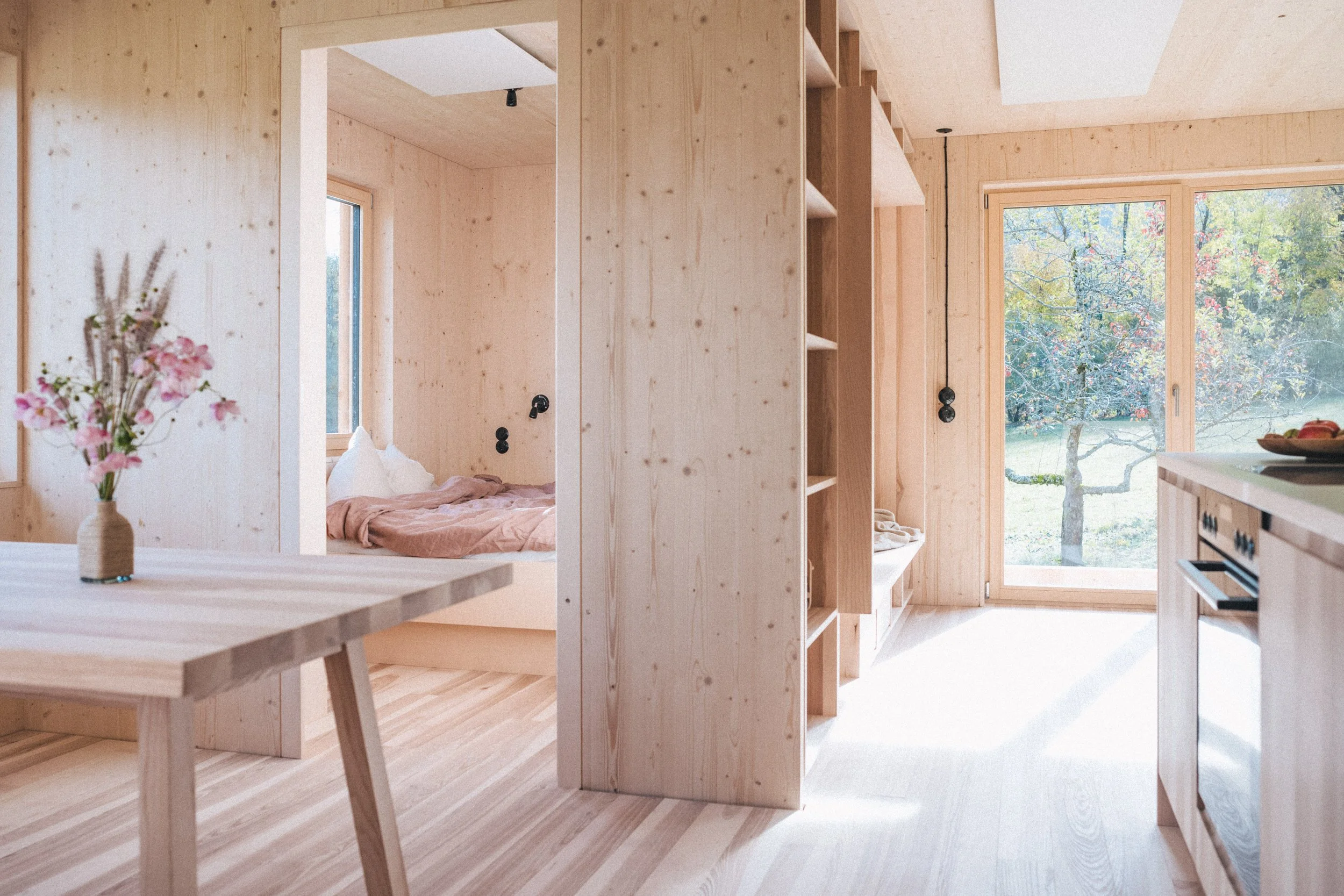 Innenansicht eines minimalistischen, hellen Holzhauses mit großen Fenstern und Ausblick auf einen Garten mit Bäumen. Teilweise sichtbares Schlafzimmer mit Bett. Essbereich mit Tisch und Blumen.