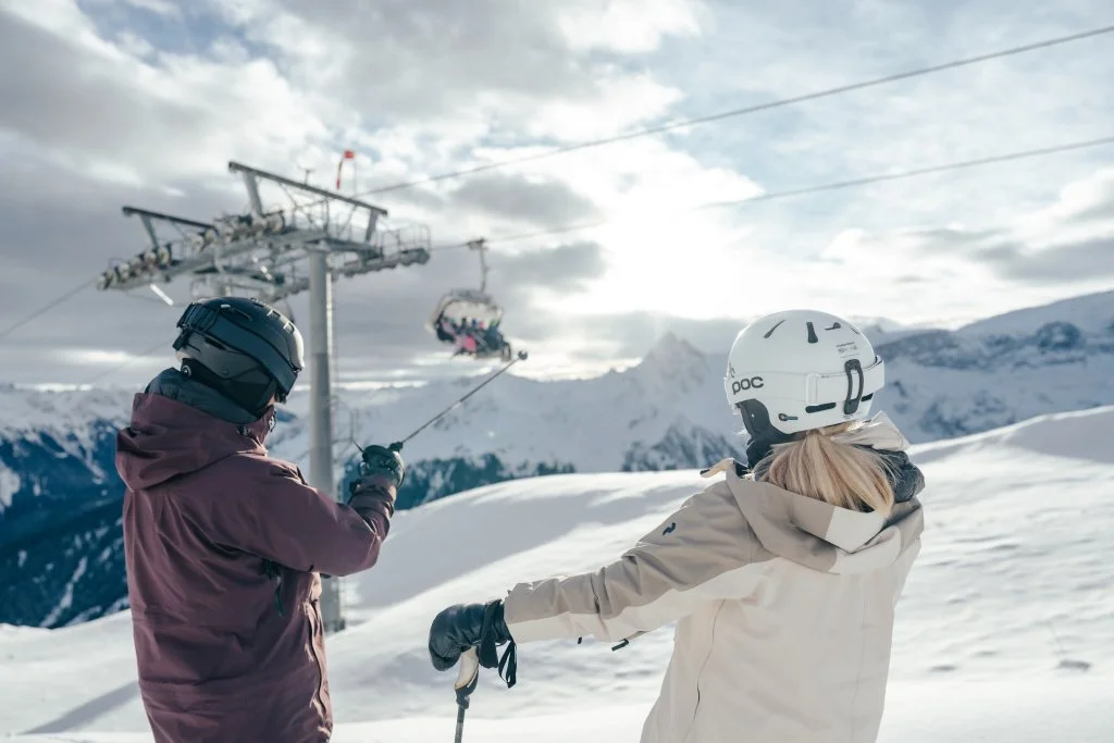Zwei Skifahrer auf einem Schneehügel mit Skilift im Hintergrund in den Bergen, eines zeigt auf den Skilift.